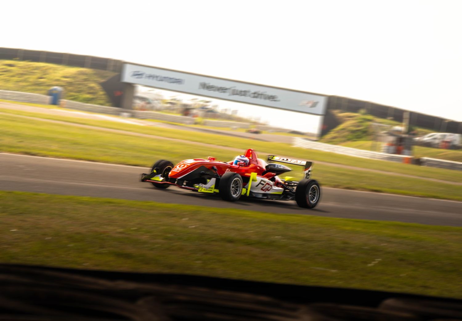 Red Formula 3 racing car accelerates on track during golden hour at a motorsport circuit.