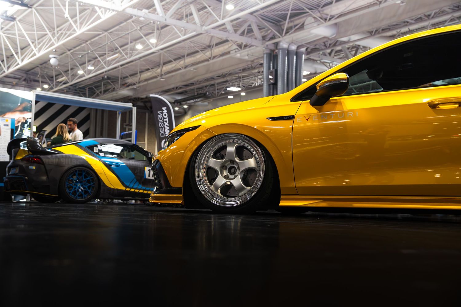 A lowered yellow Ford Focus ST and blue race car on display at an indoor car show.