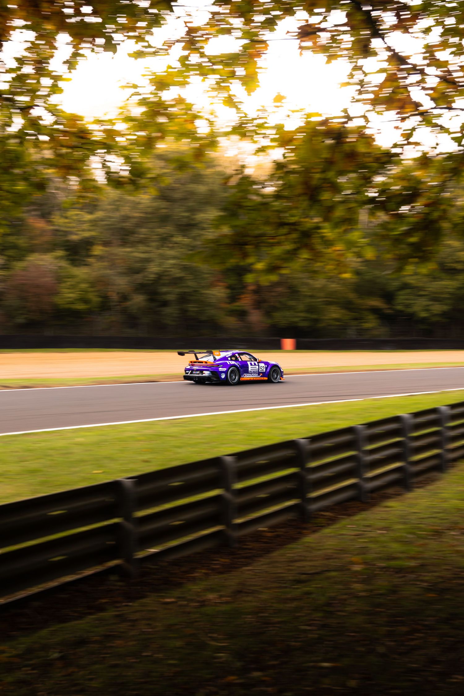 A purple GT race car speeds past autumn trees along a fenced circuit at golden hour.