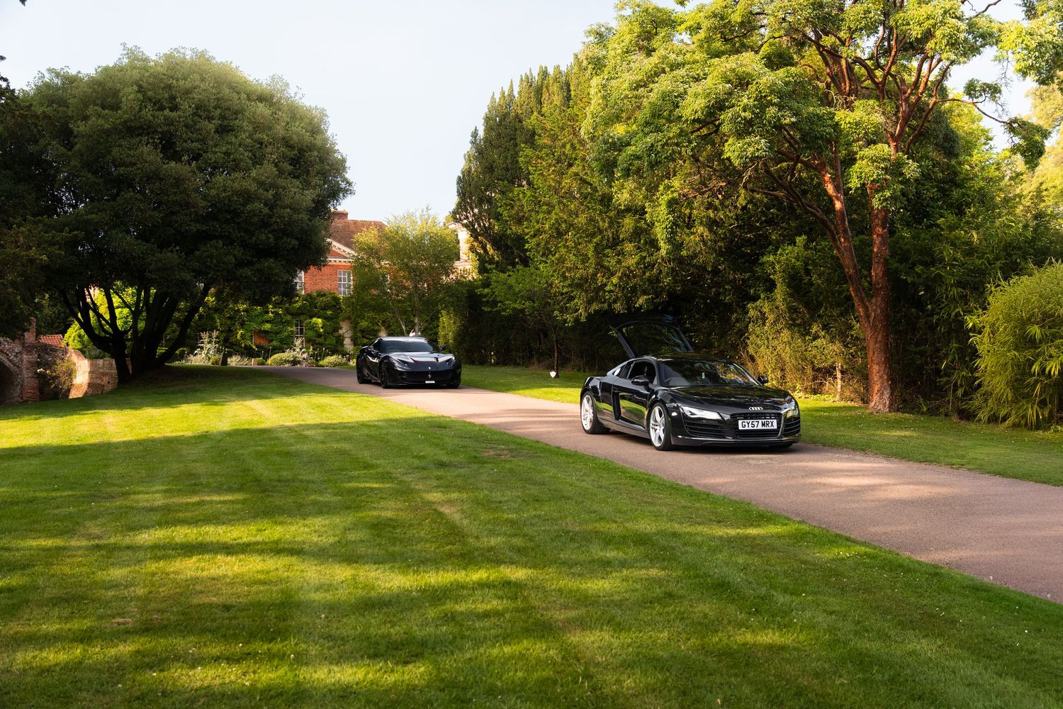 Two black luxury sports cars parked on a gravel driveway surrounded by lush green lawn and trees.