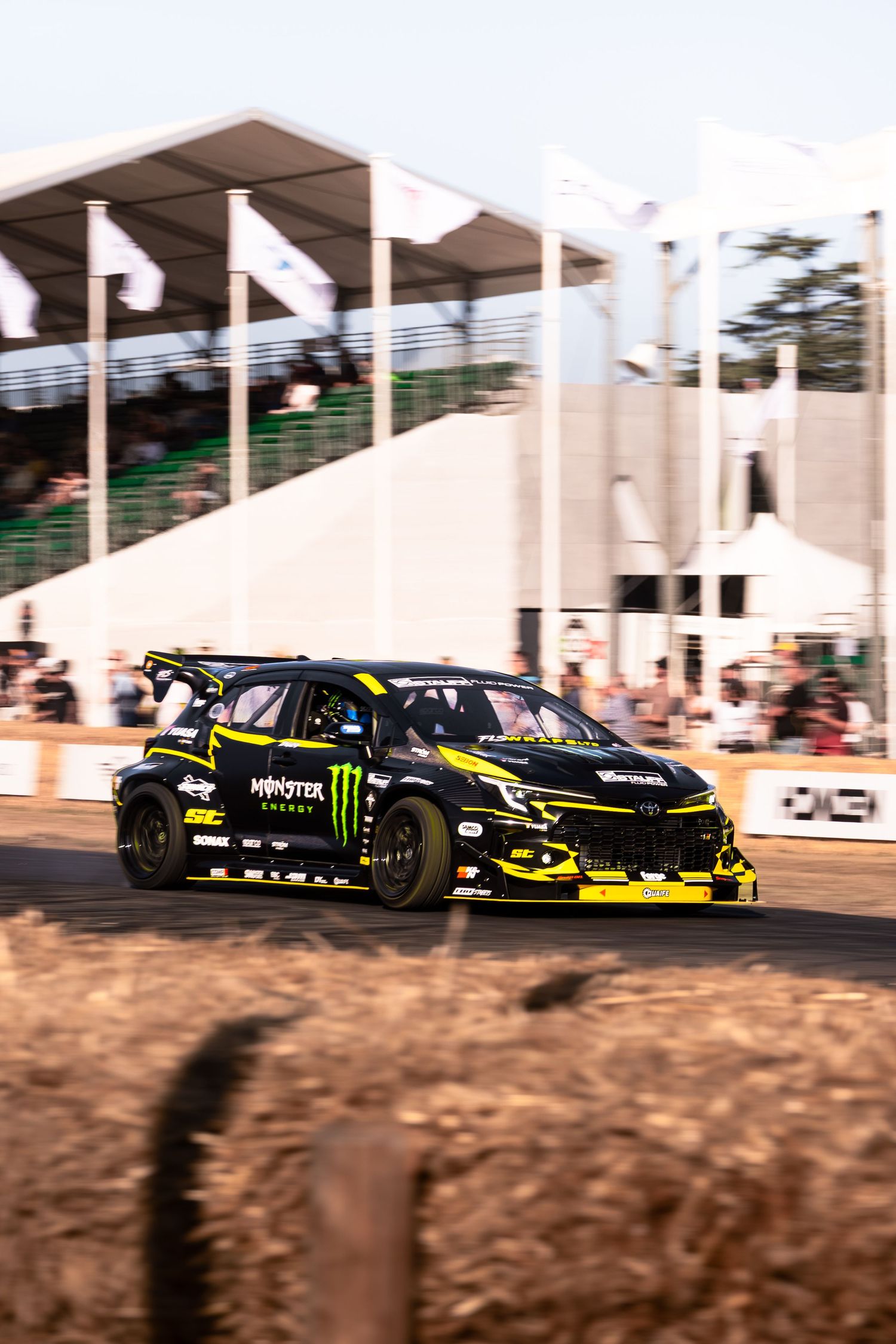 Monster Energy black rally car racing on dirt track at outdoor motorsport event with grandstand in background.