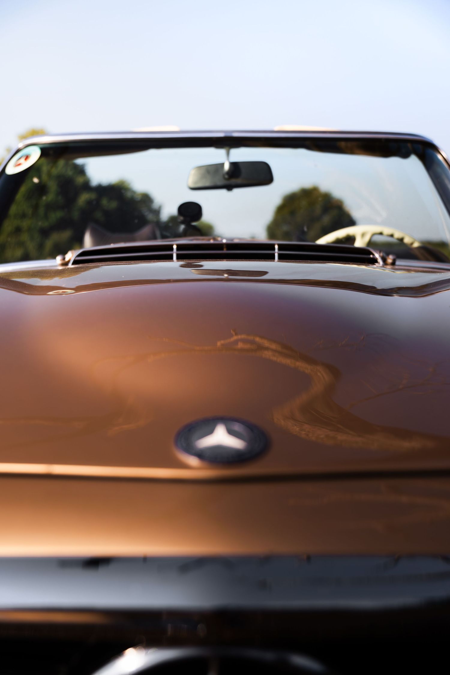 Vintage bronze Mercedes-Benz convertible car hood with iconic three-pointed star emblem under clear blue sky.