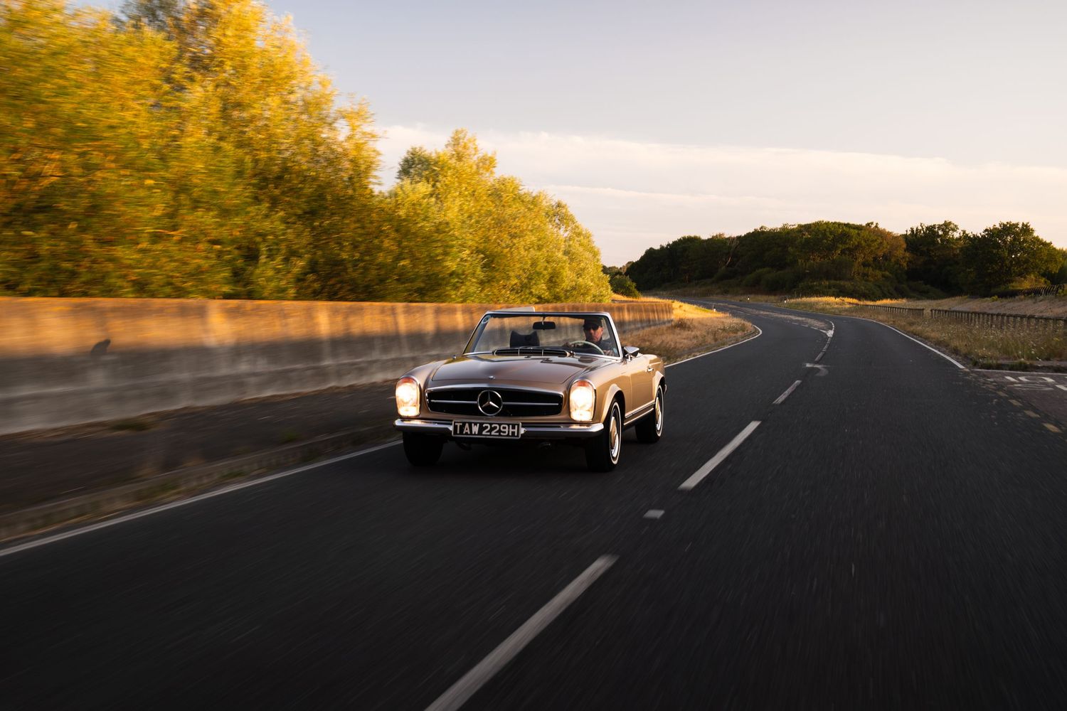Vintage Mercedes-Benz convertible cruising on an open road at golden hour, surrounded by lush autumn trees.