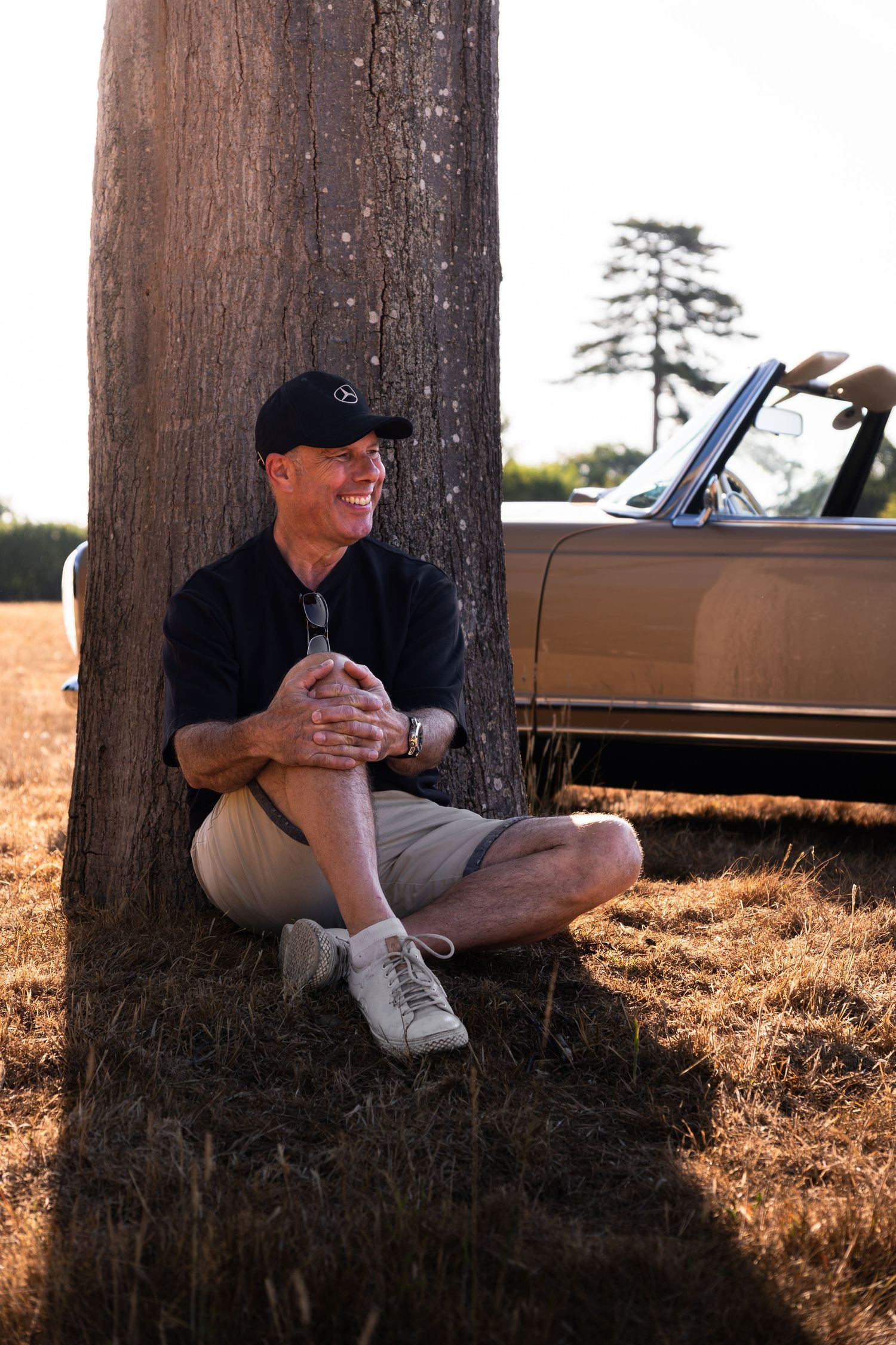 Man in black cap and shorts sits against a tree beside a vintage convertible car in a sunny field.