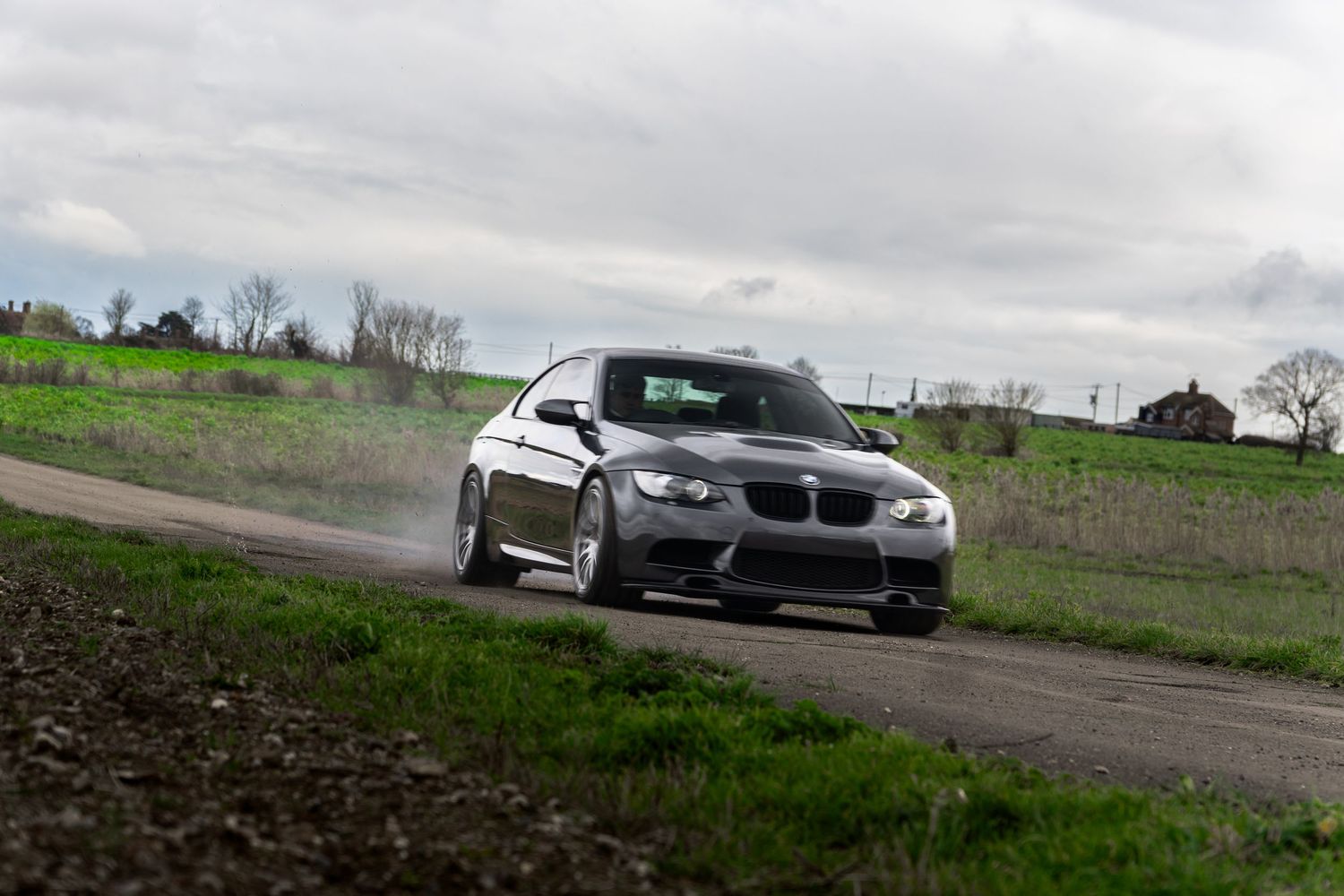 Silver BMW M3 E92 coupe cornering on a wet rural road with green grass and countryside in the background.