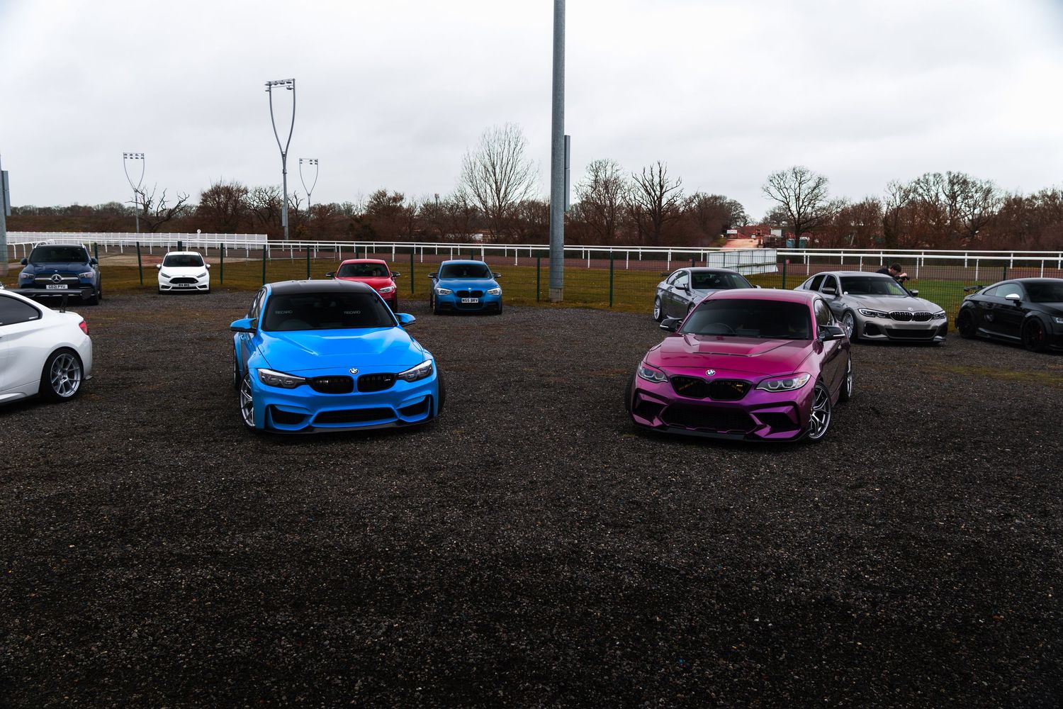 Blue and pink widebody BMW M3 and M2 cars parked at an outdoor car show event.