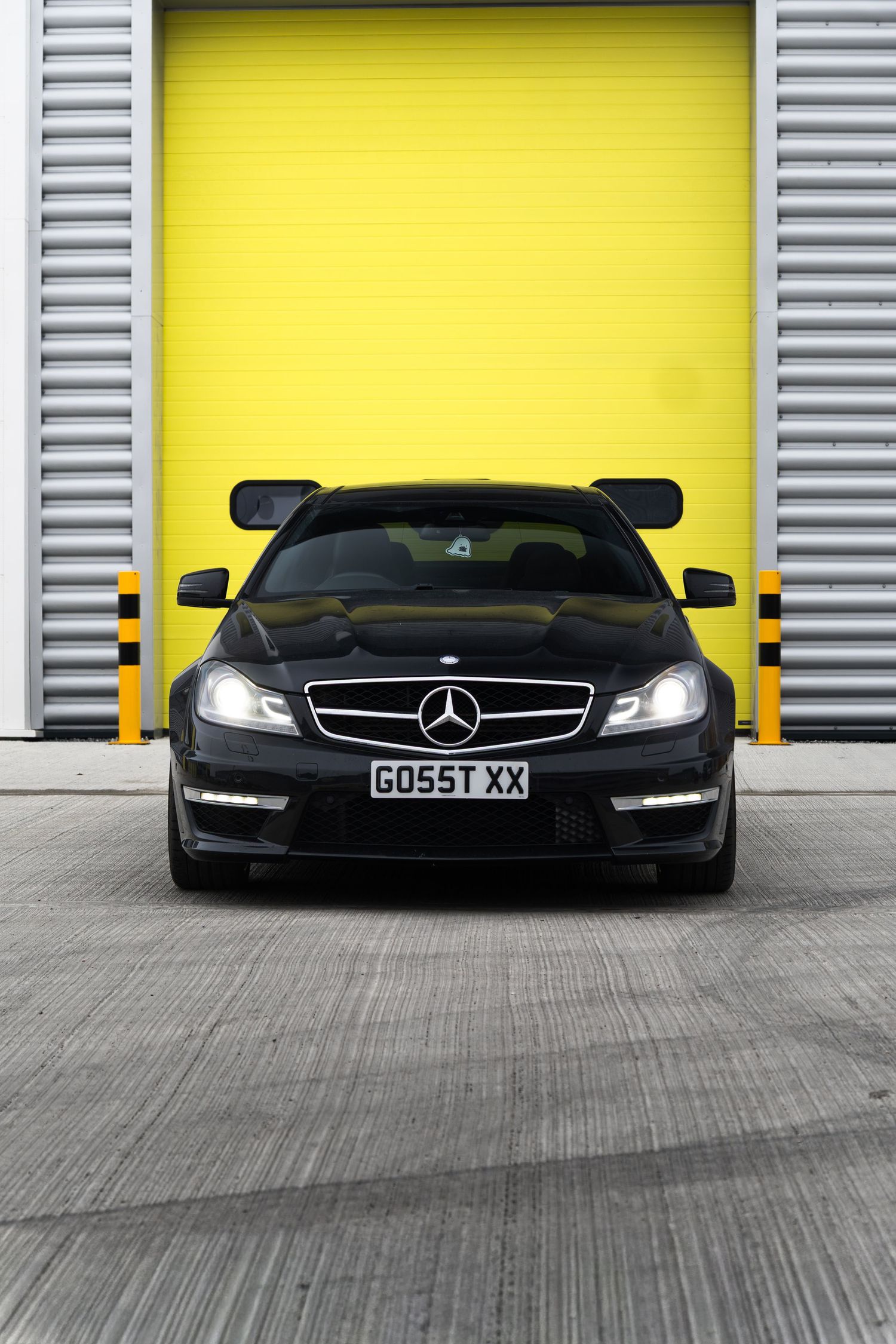 Black Mercedes-Benz C-Class AMG coupe with GO55T XX plate parked directly in front of bright yellow garage door.