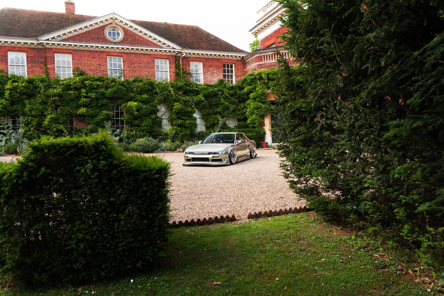 Silver sports car parked on gravel driveway of grand red brick manor house surrounded by lush green hedges and ivy.