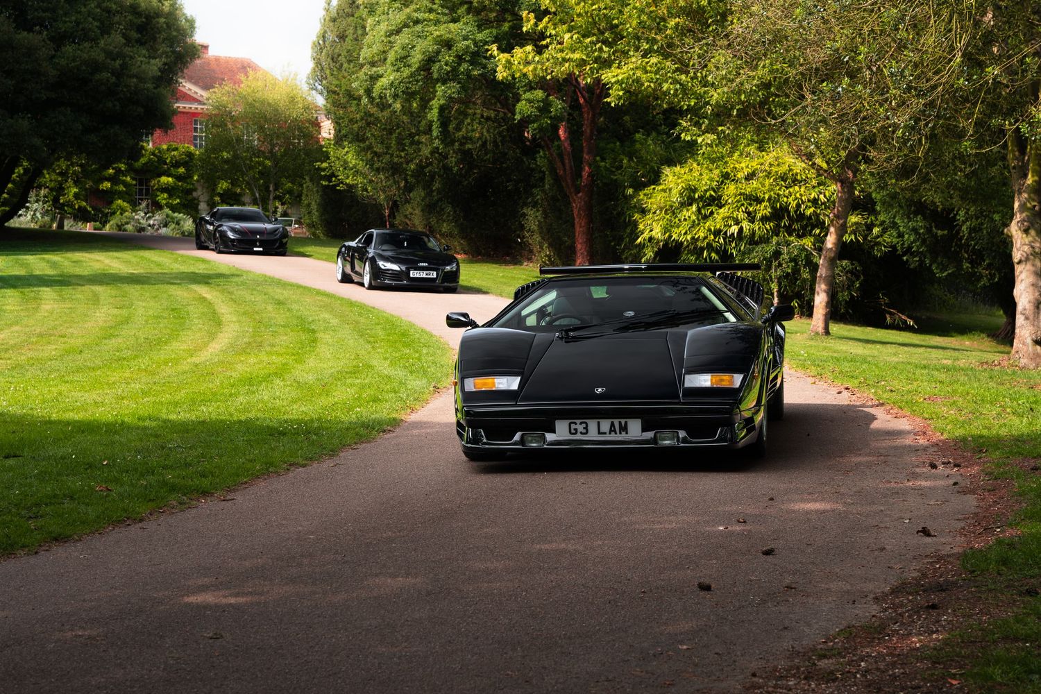 Black Lamborghini Countach driving on a tree-lined path in a lush green park setting.