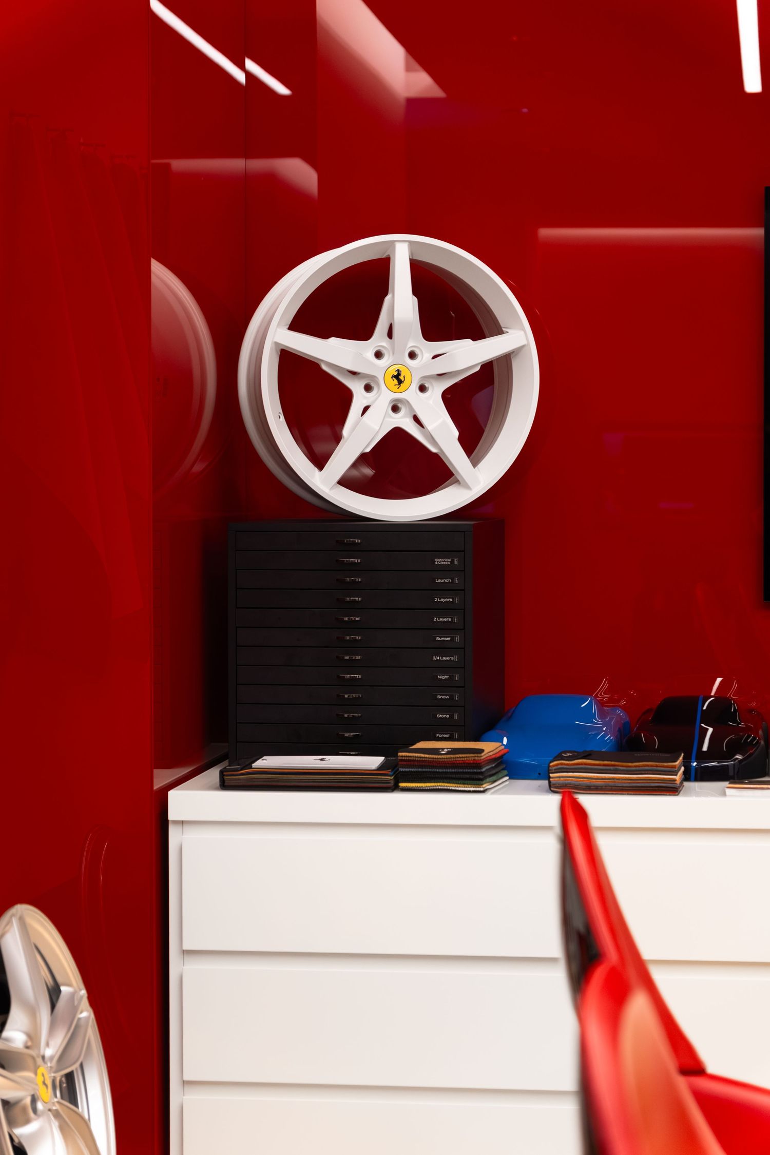 White Ferrari five-spoke alloy wheel displayed on a white dresser against a bold red wall in a Ferrari showroom.