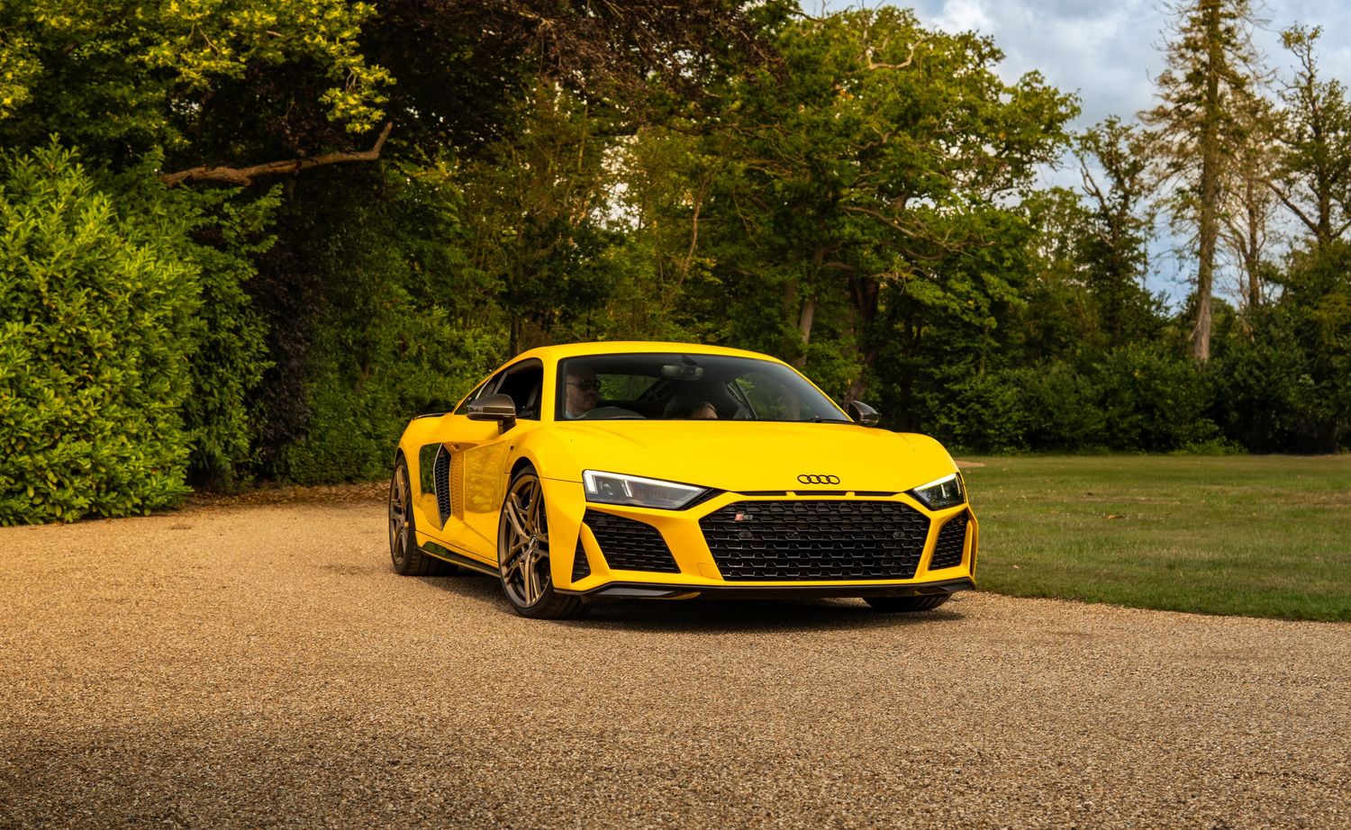 Yellow Audi R8 supercar parked on a gravel driveway surrounded by lush green trees.