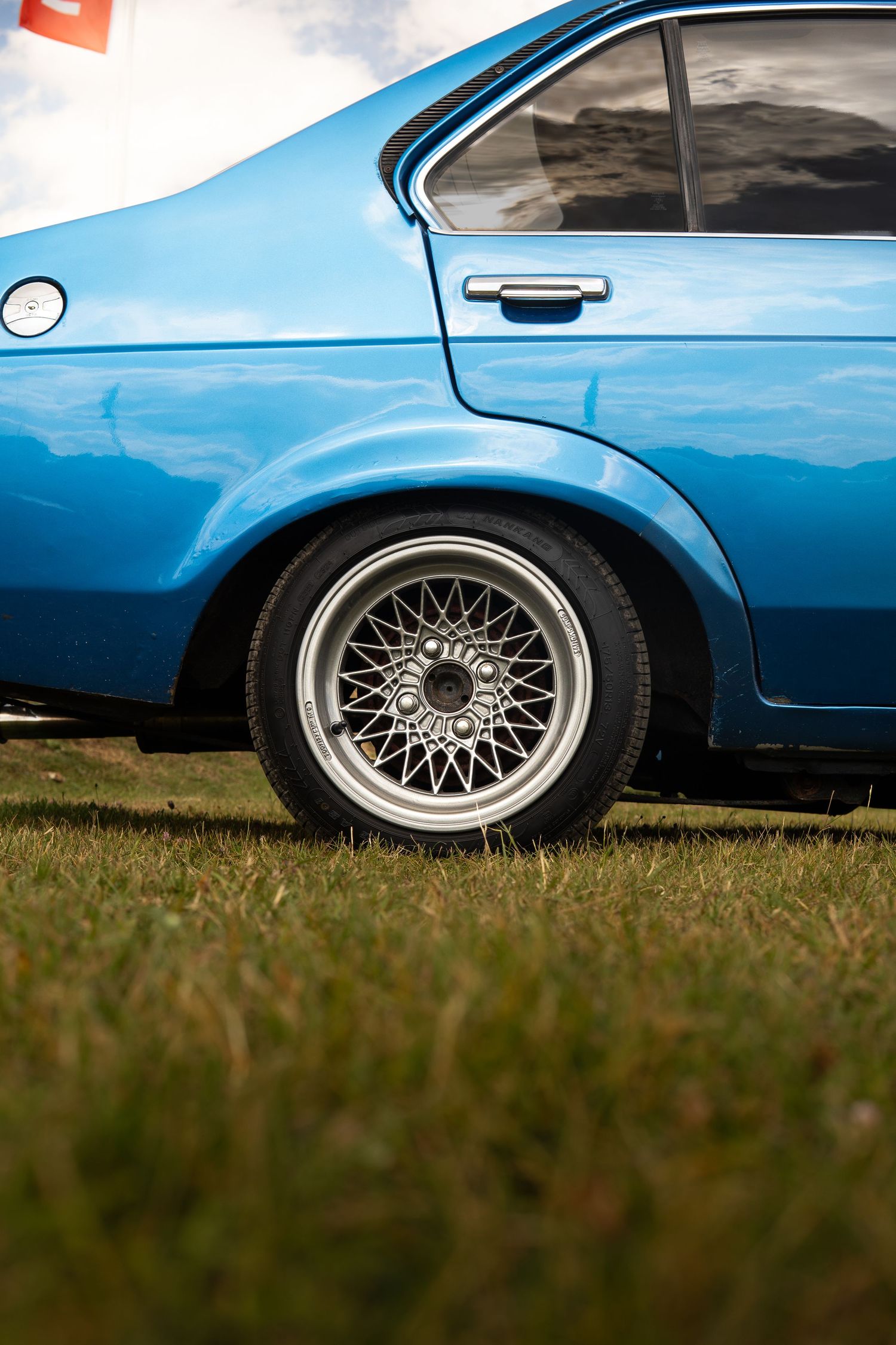Side profile of a classic blue coupe with silver mesh alloy wheel parked on grass.