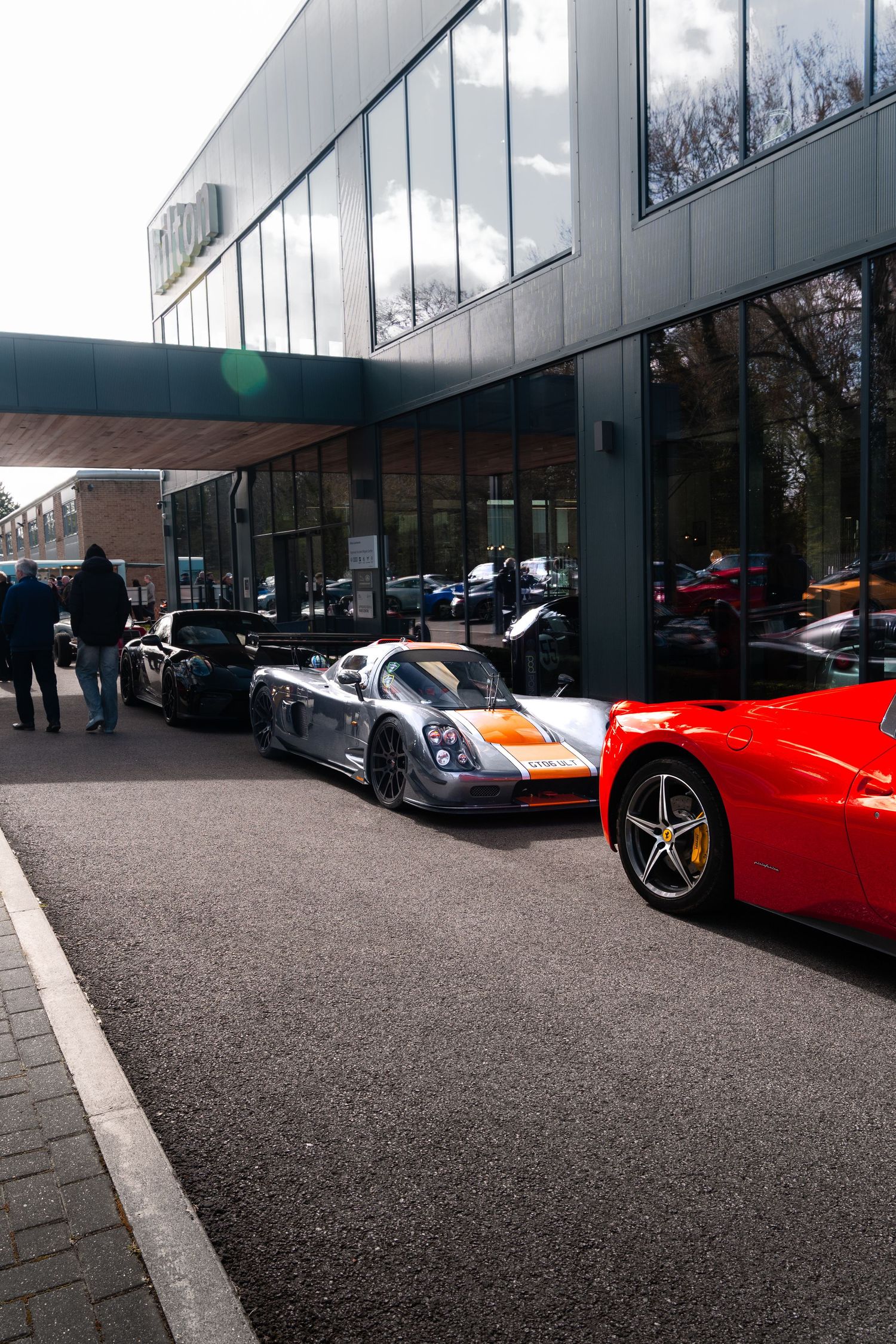 Mosler MT900 race car and red Ferrari parked outside modern glass building.