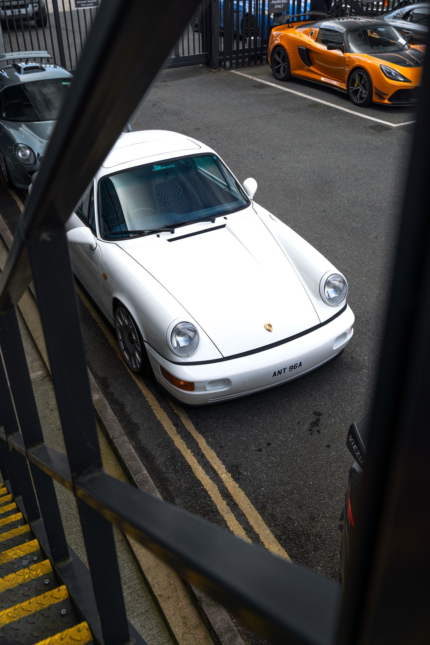 White Porsche 964 911 viewed from above through metal staircase with yellow Lotus in background.