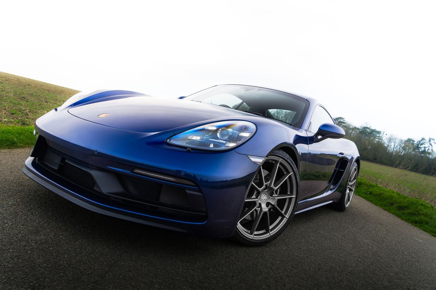 Low-angle front shot of blue Porsche 718 Cayman on a winding road with green hillside.