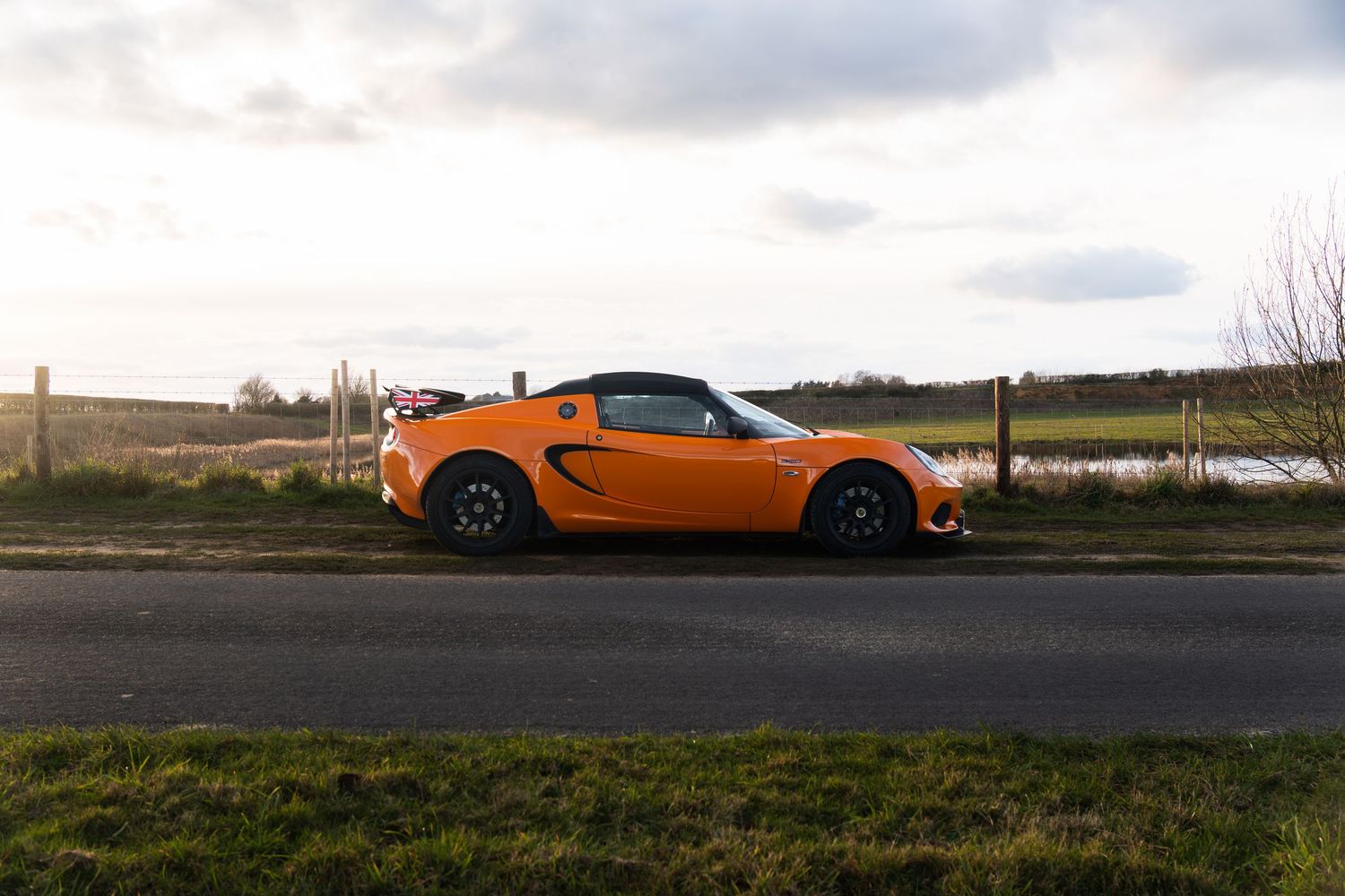 Bright orange Lotus Elise sports car parked on rural countryside road beside green fields.