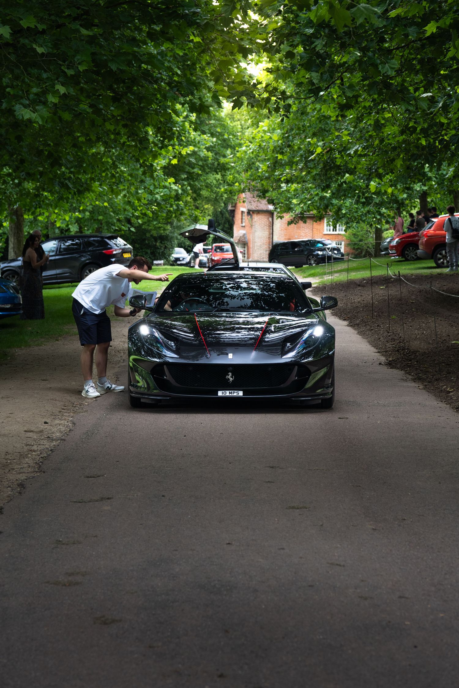 A dark Ferrari sports car parked on a tree-lined path, with a person leaning against it at an outdoor car show.