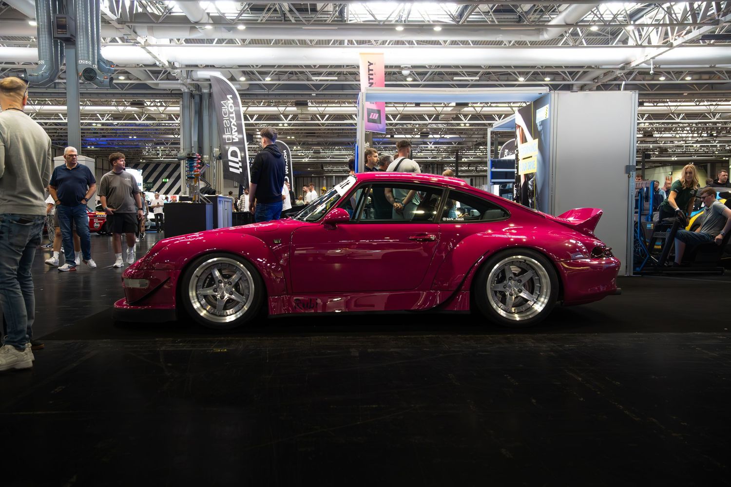 A pink RWB wide-body Porsche 993 on display at an indoor automotive exhibition with spectators.