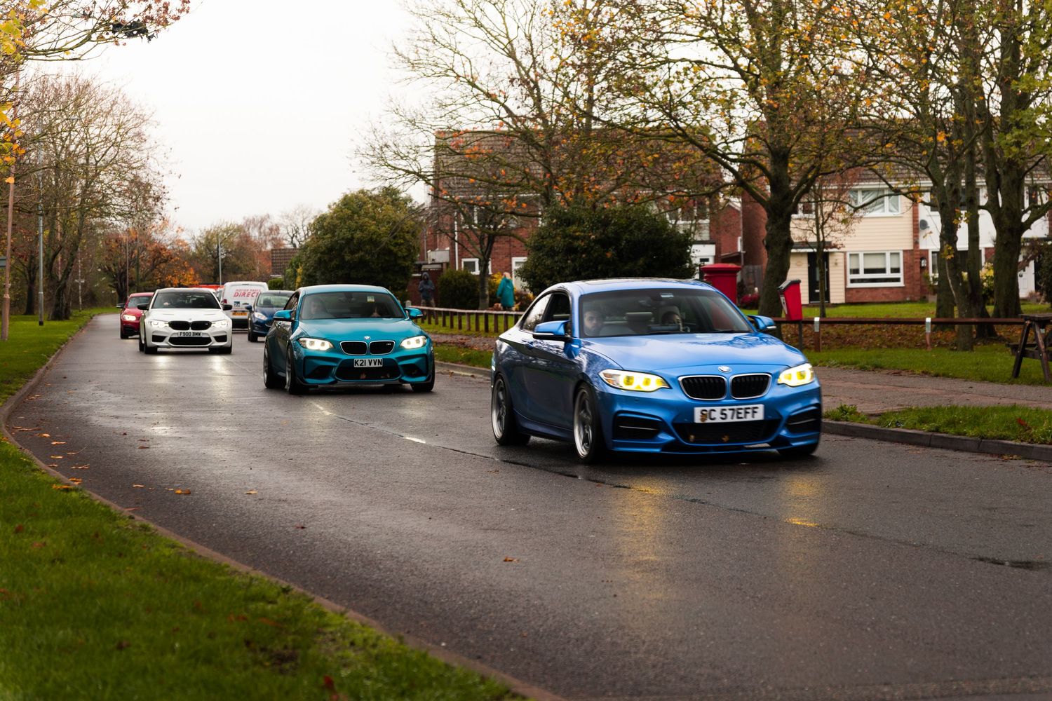 Blue BMW 2 Series leading a convoy of modified cars on a wet road.
