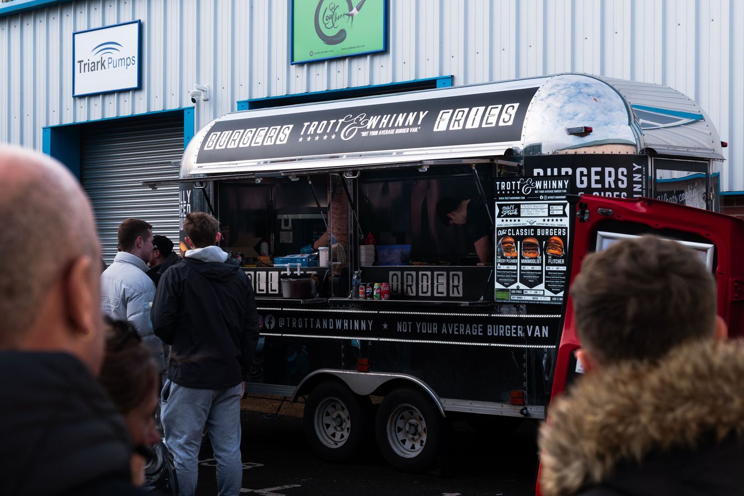 A food truck serving burgers attracts a crowd at an outdoor car meet event.