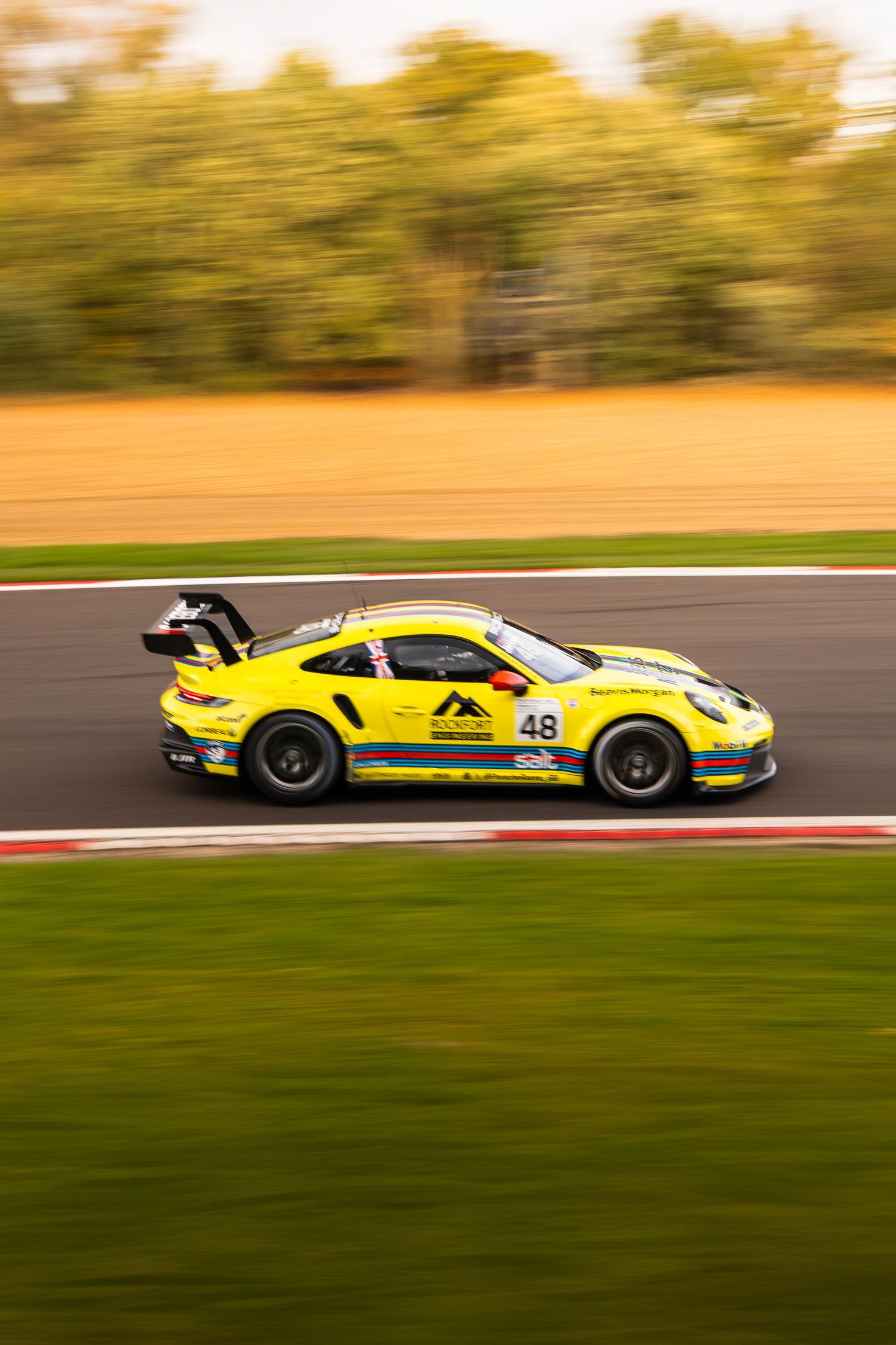 Yellow Porsche 911 GT3 number 48 racing on a circuit with golden autumn trees and green grass.