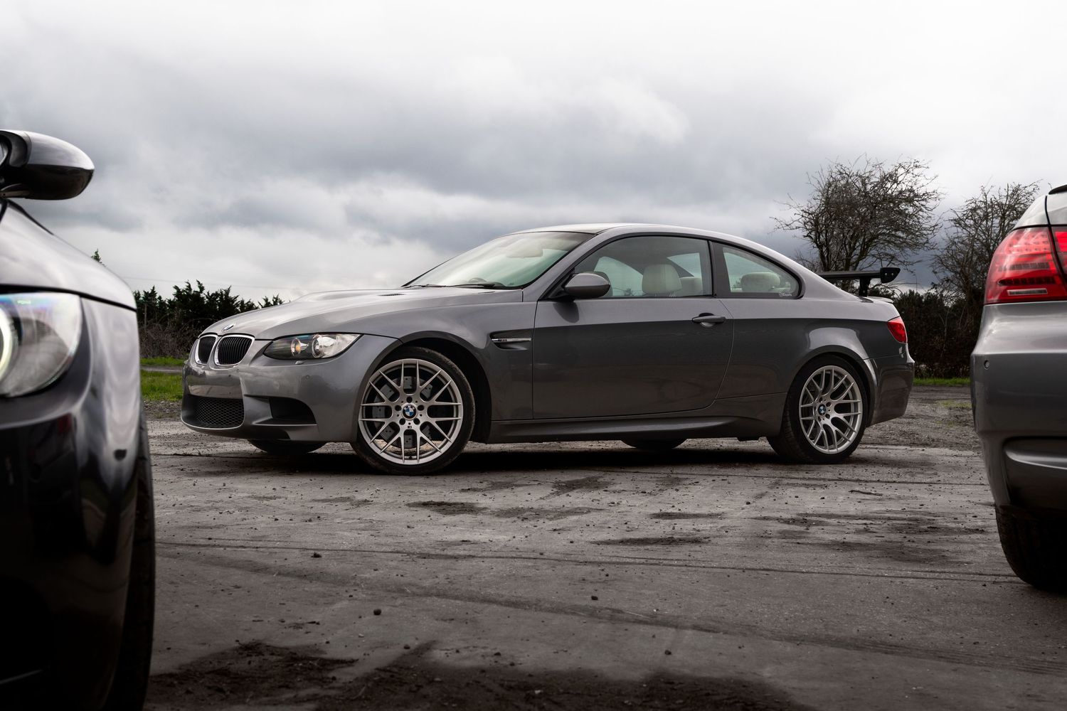 Side profile of a grey BMW M3 coupe with silver alloy wheels parked on wet tarmac.