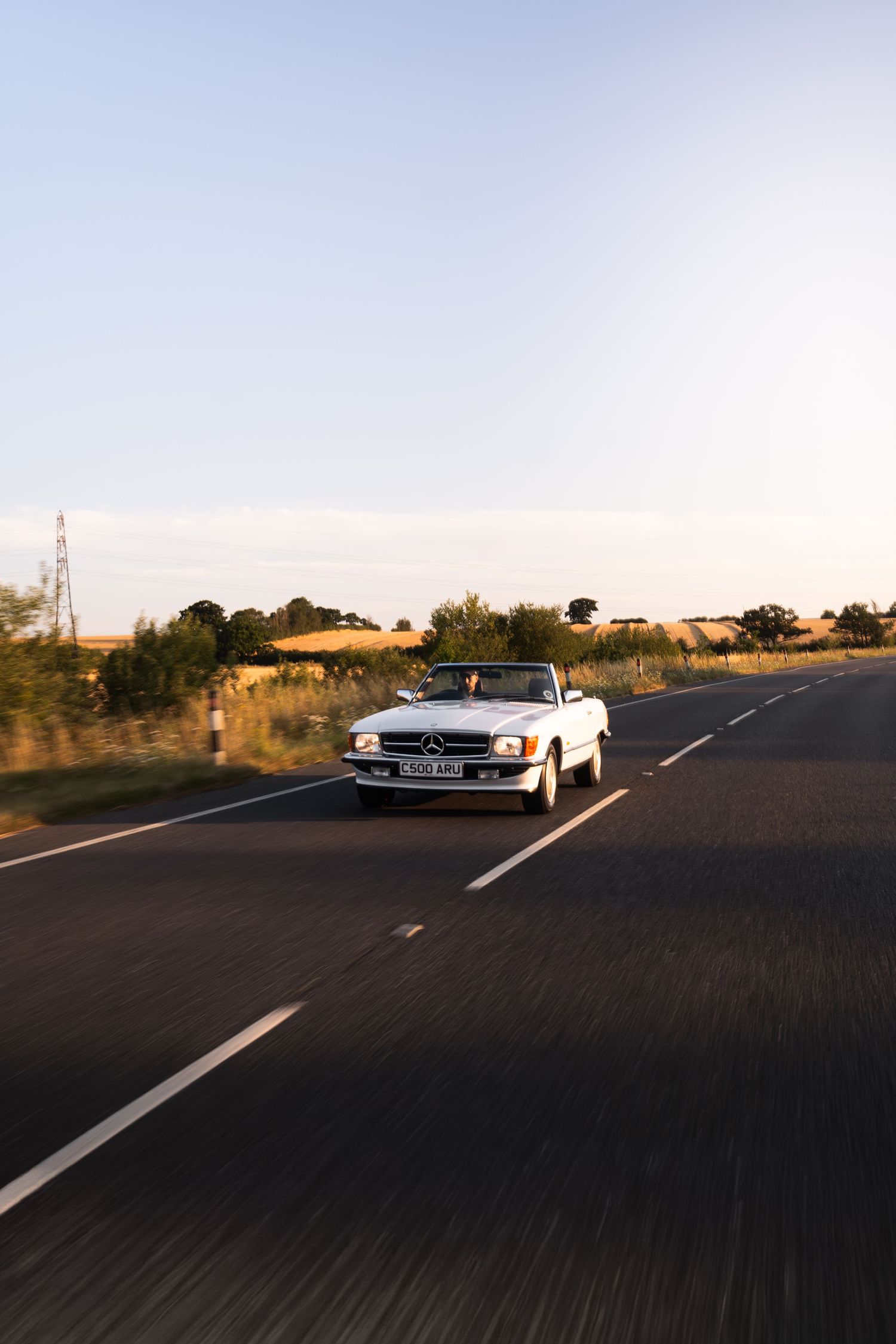 White Mercedes-Benz convertible driving on a rural road at golden hour with clear skies and fields in the background.