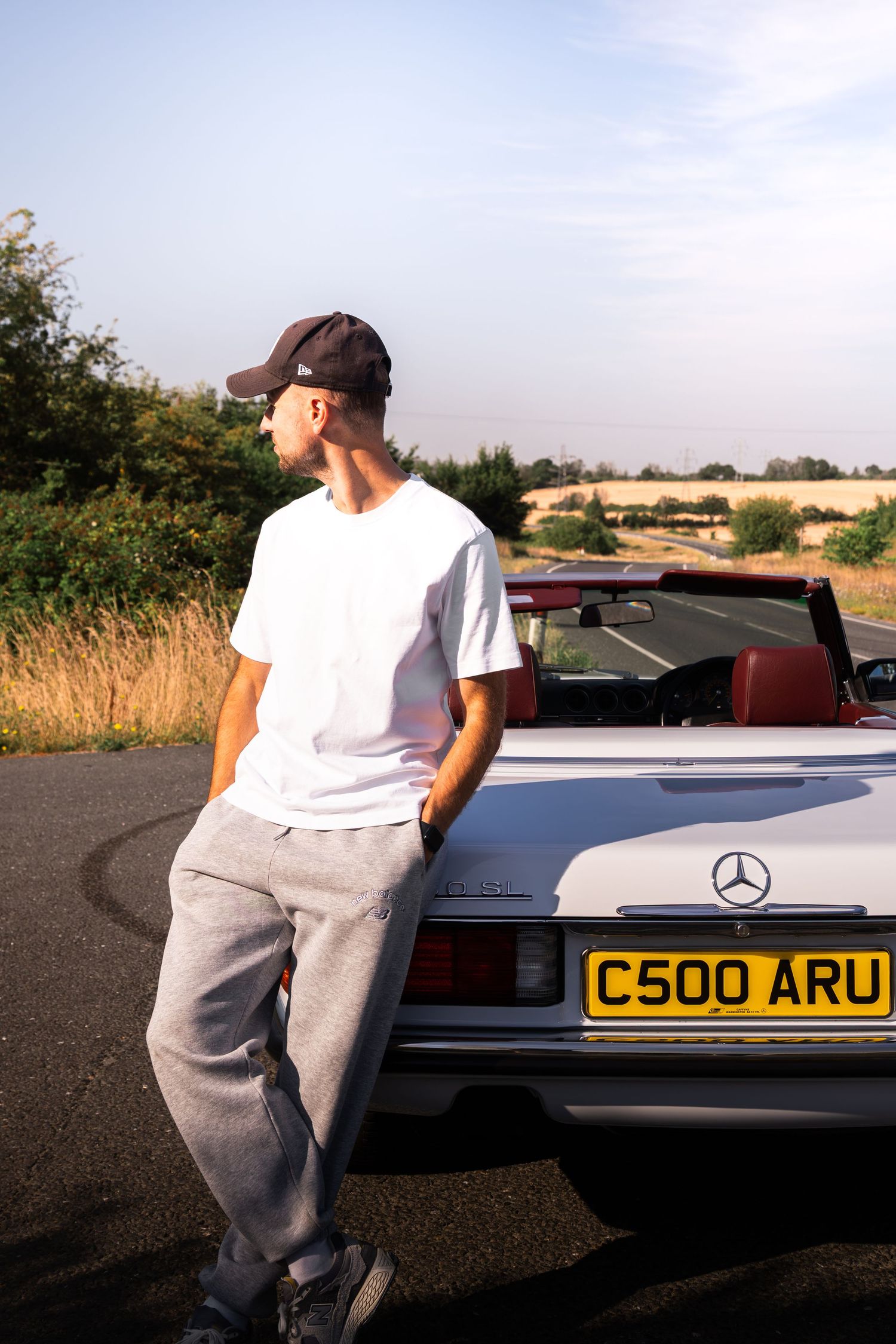 Young man in white tee and grey sweats leans against a silver Mercedes convertible with UK plate C500 ARU on a rural road.
