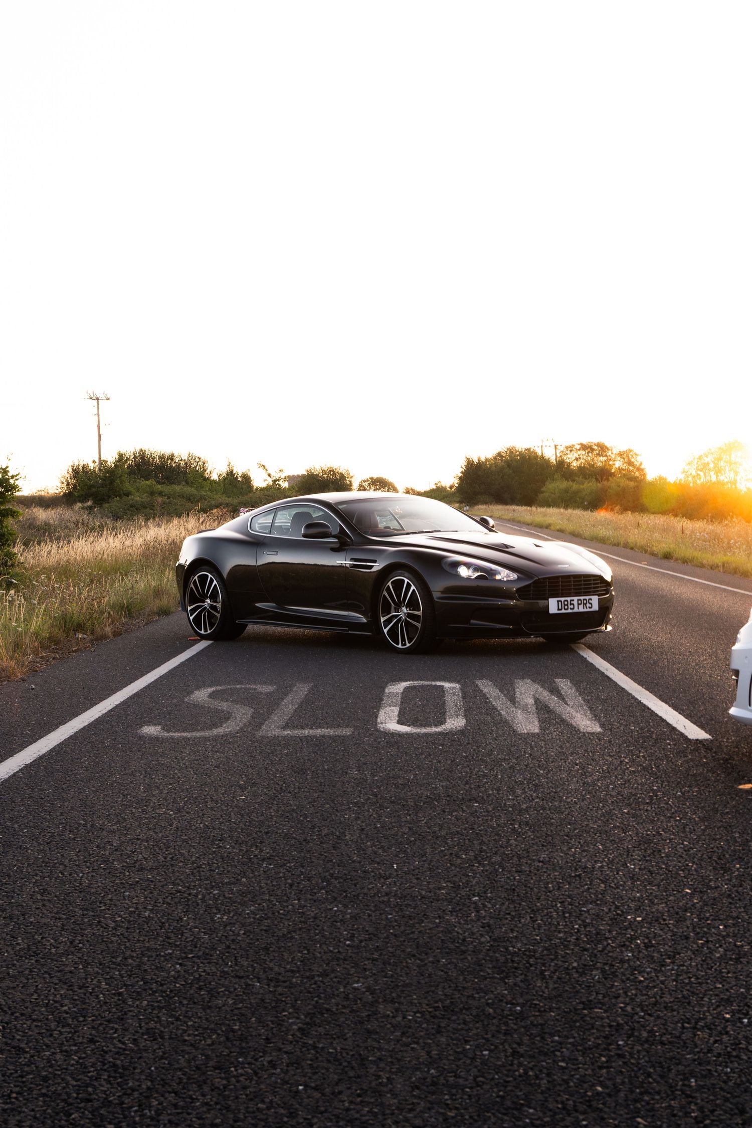 Black Aston Martin DBS parked on a road with SLOW marking at golden sunset, surrounded by countryside.
