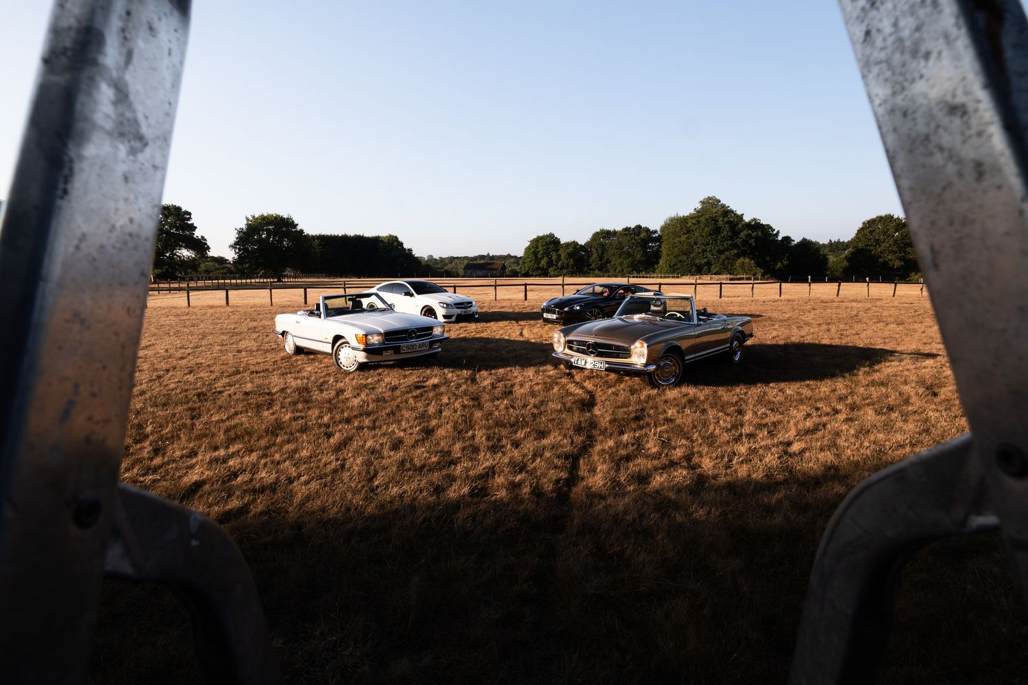 Classic American muscle cars parked in a golden field, viewed through a metal frame gate at sunset.
