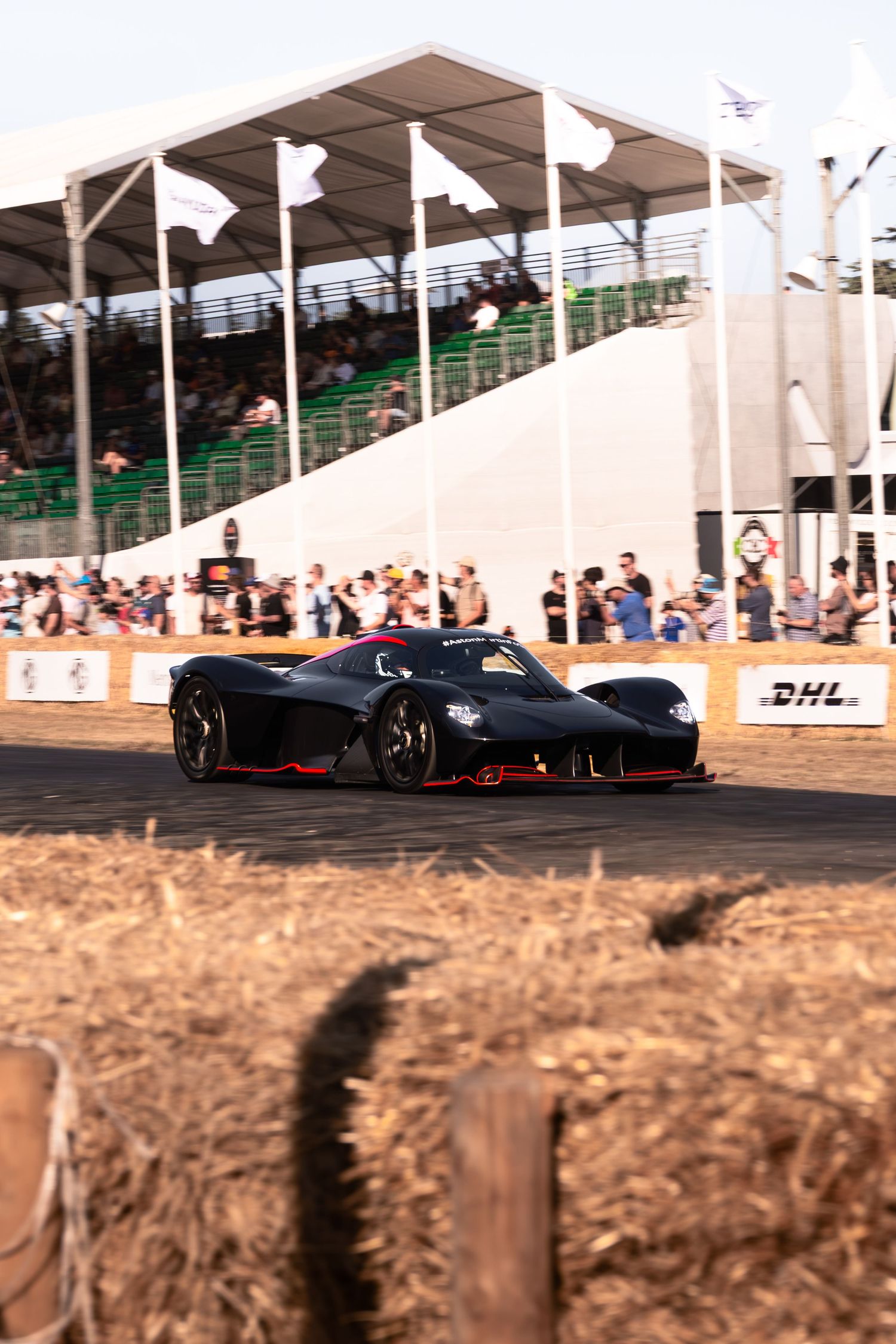 Black Aston Martin Valkyrie hypercar racing at Goodwood Festival of Speed with spectators and DHL signage.