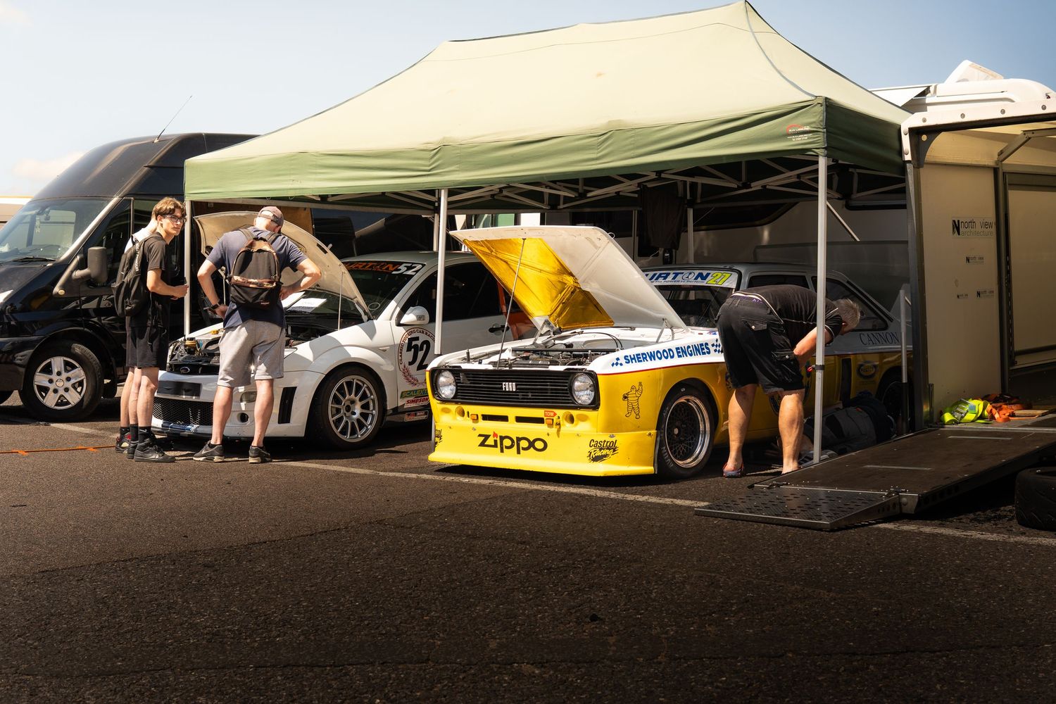 Yellow Zippo sponsored BMW race car with hood open being serviced under green tent.