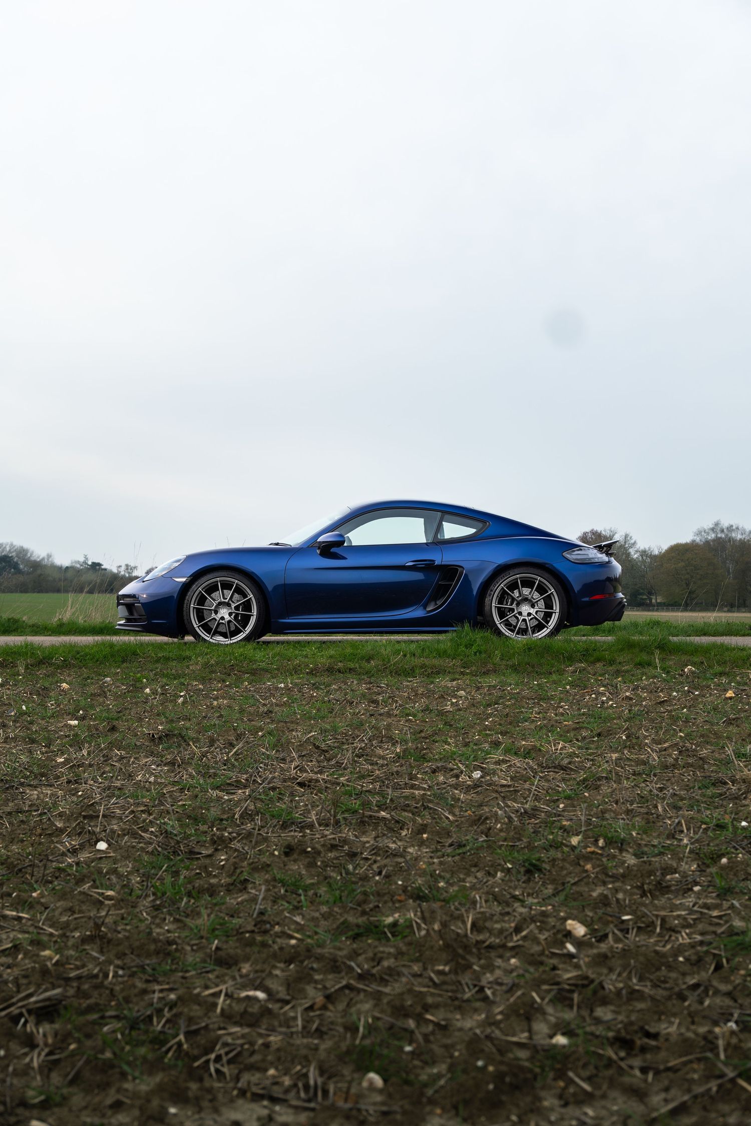 Blue Porsche 718 Cayman parked on a rural road with green fields and overcast sky in background.