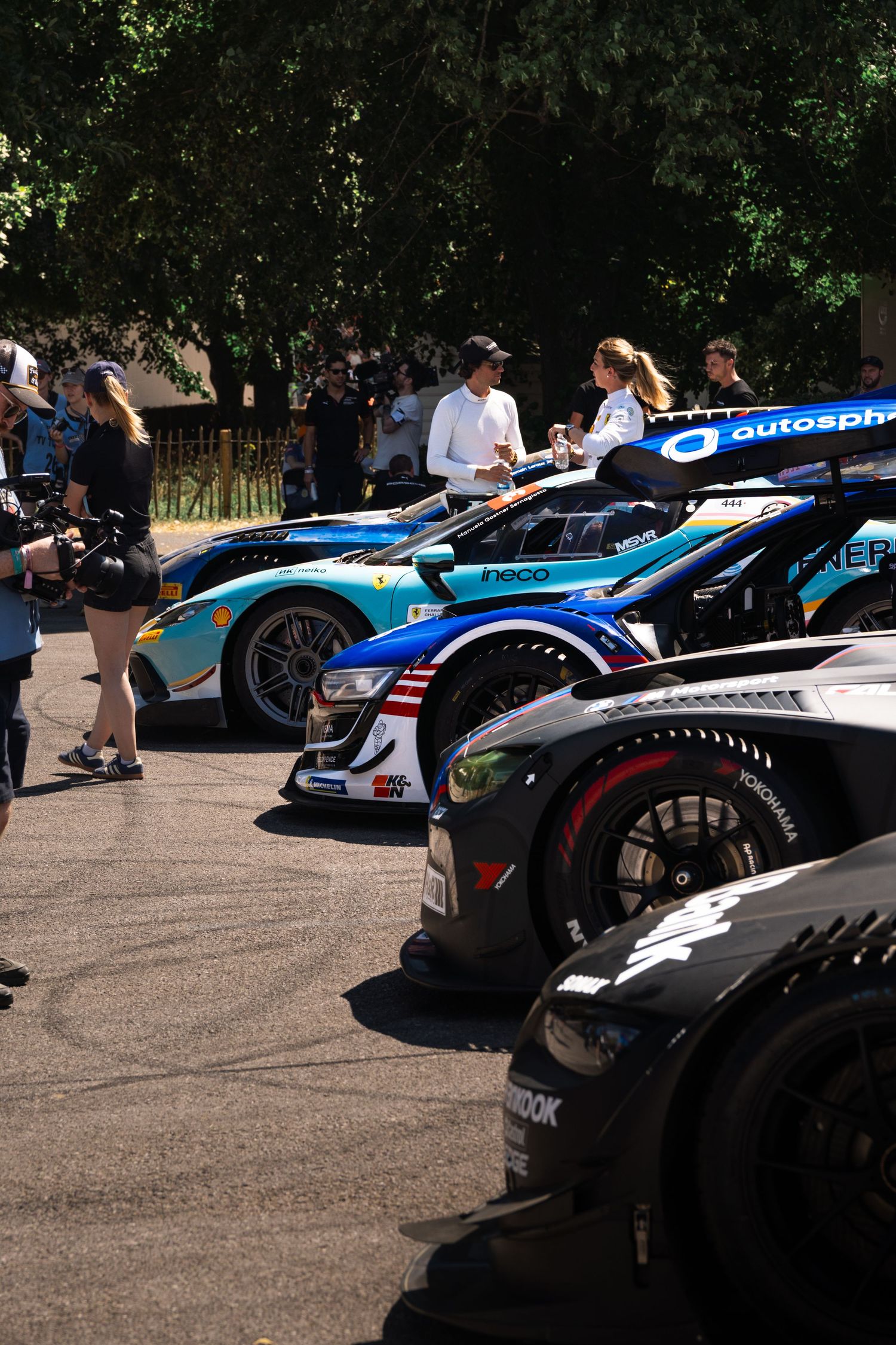 Row of GT racing cars lined up at a motorsport event, with spectators and camera crew visible in the background.