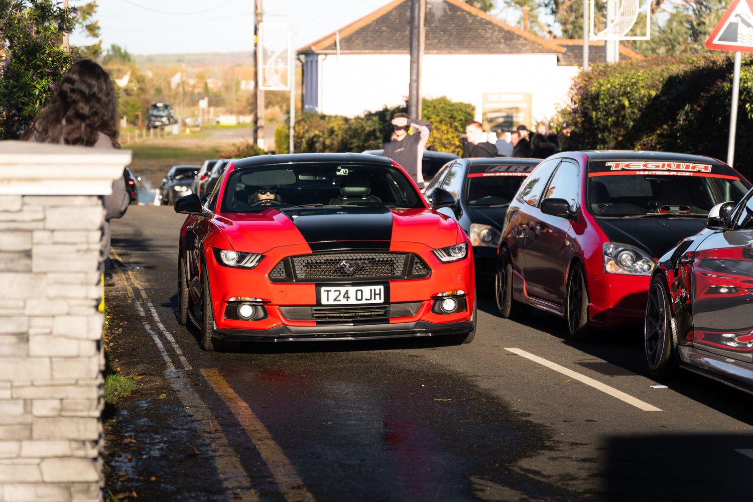 Red Ford Mustang with black racing stripes driving through a village street car meet.