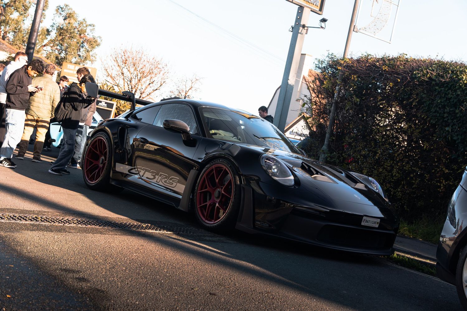 Black Porsche 911 GT3 with red wheels at an outdoor car meet on a sunny day.