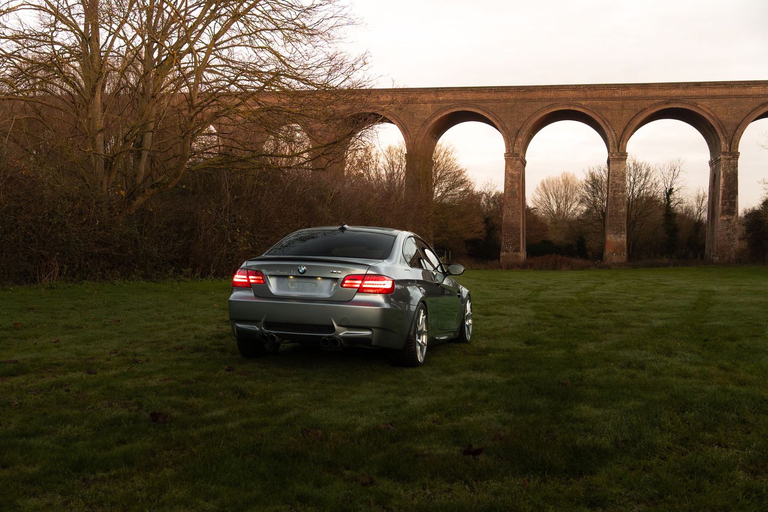 Grey BMW M3 coupe viewed from rear parked on grass before tall brick viaduct.