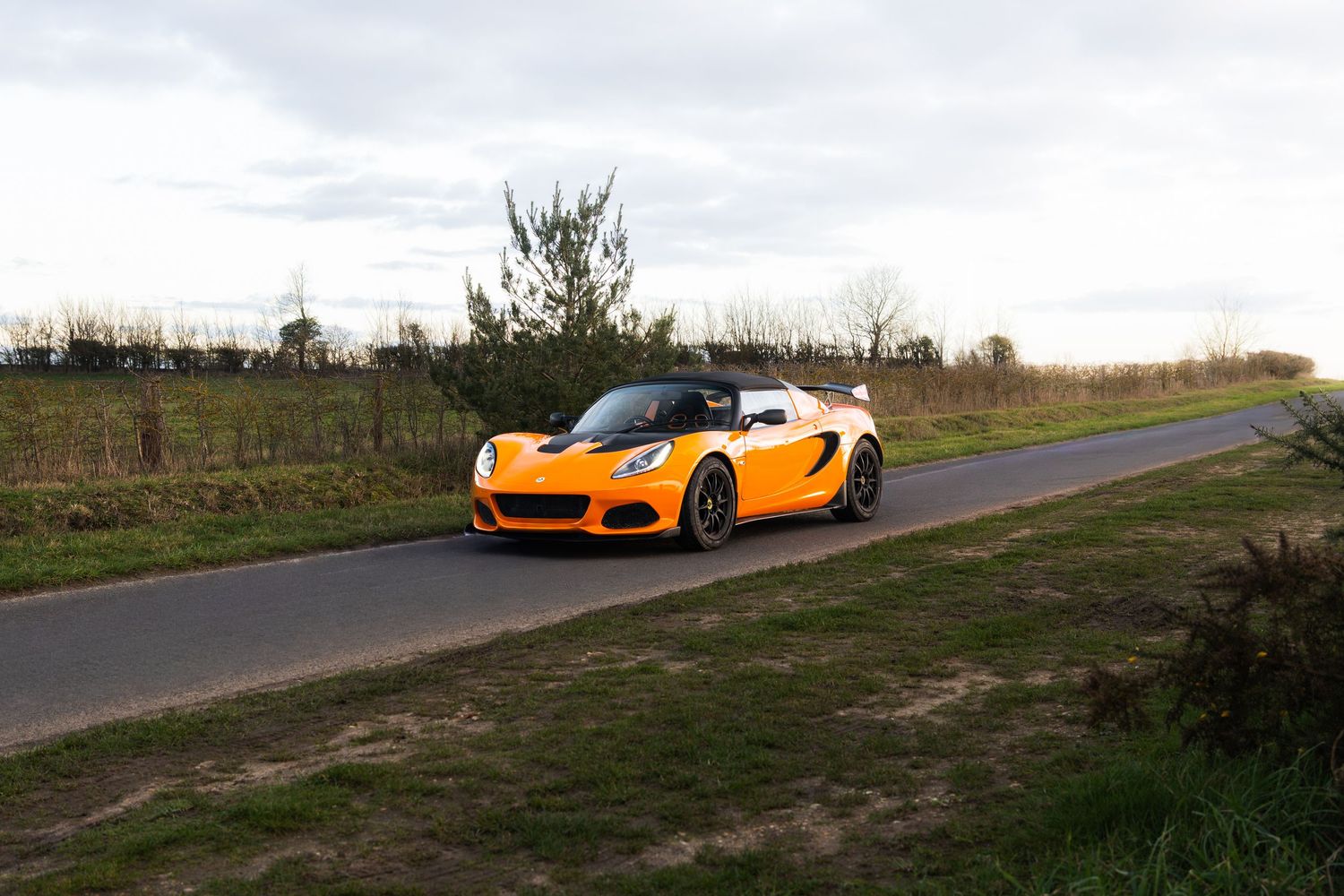 Orange Lotus Elise sports car driving on a narrow country road surrounded by green fields.