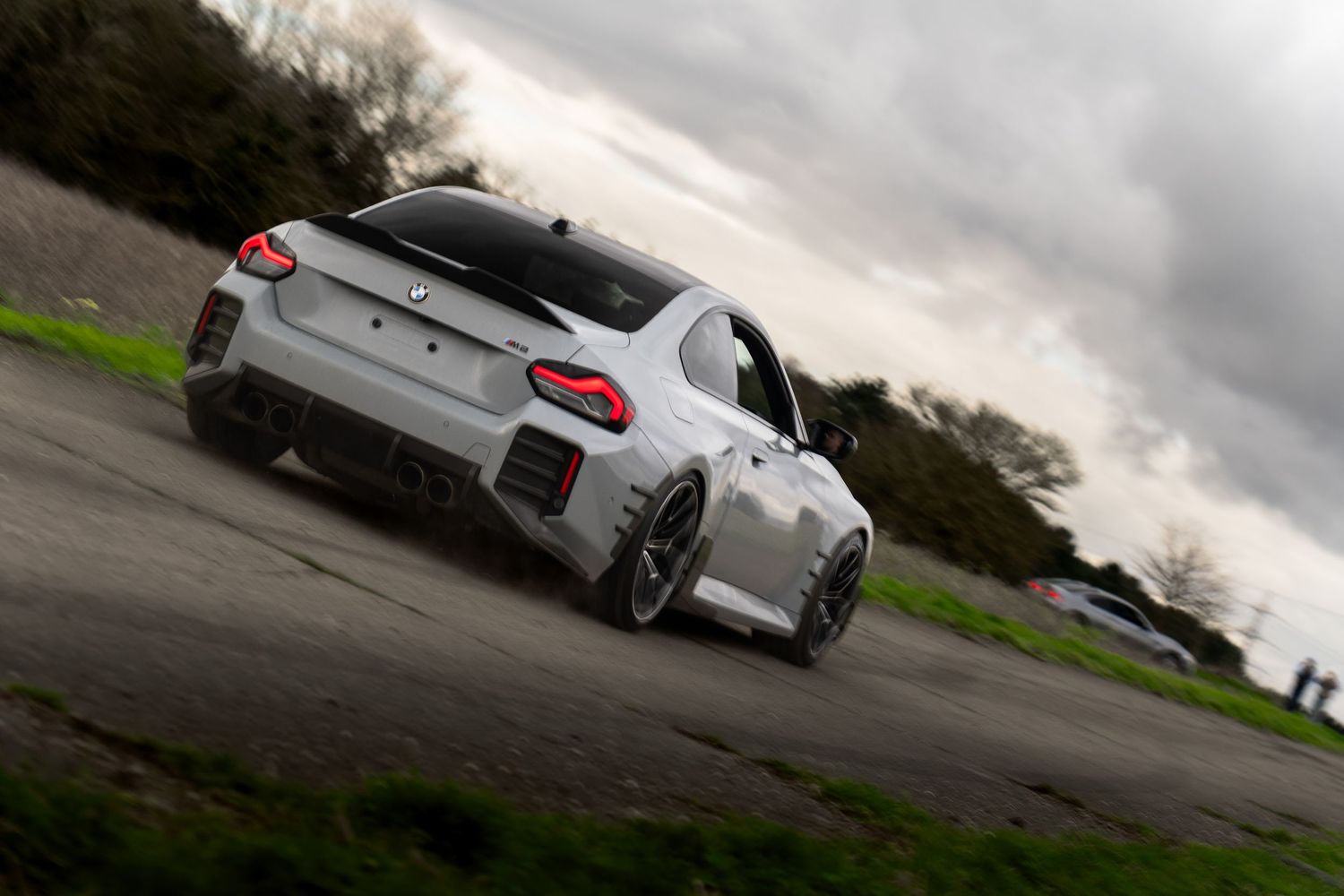 Rear view of a white BMW M2 with carbon fiber spoiler and wide body kit on a rural road under dramatic clouds.