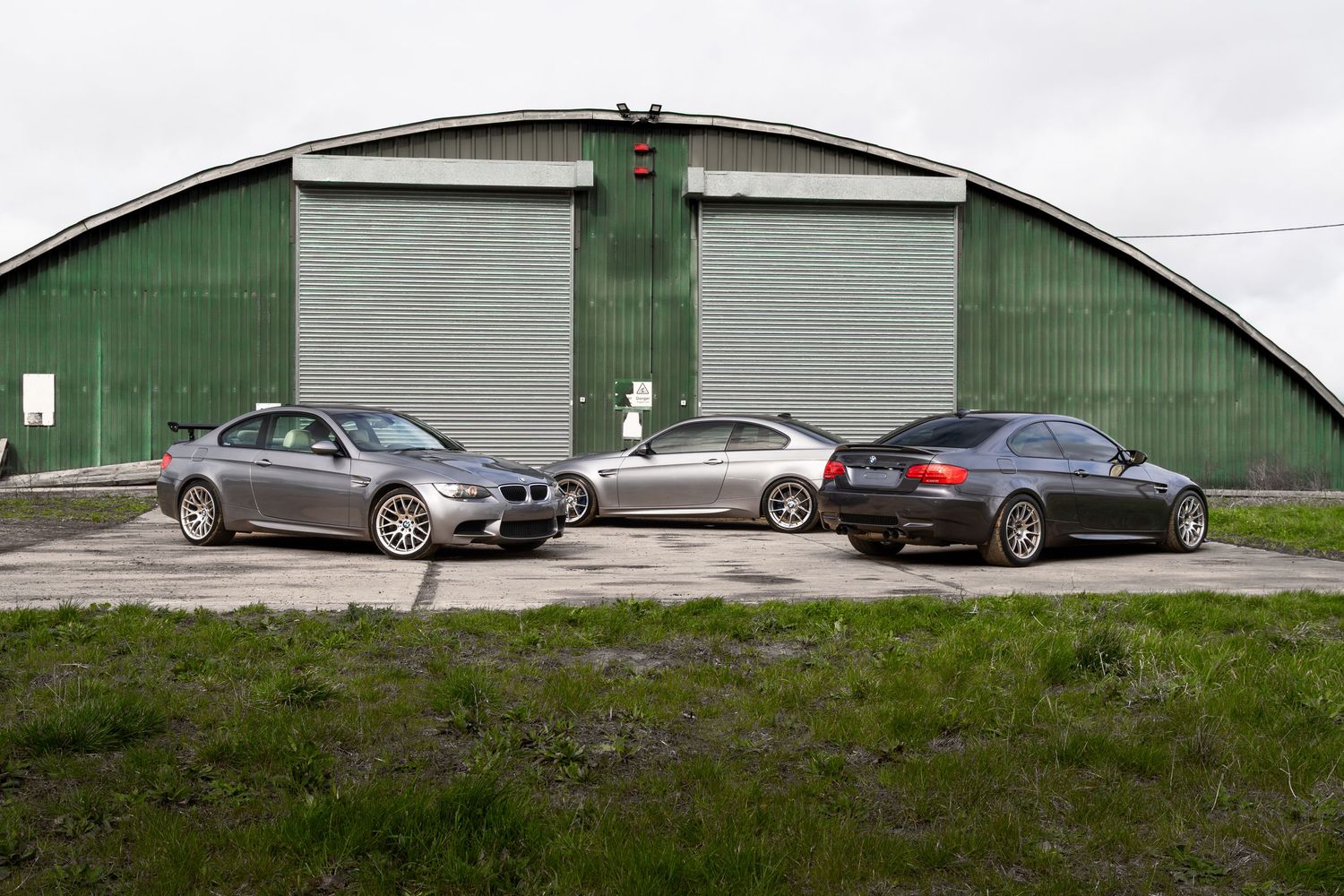 Three BMW M3 vehicles lined up in profile view outside a large green arched metal hangar.