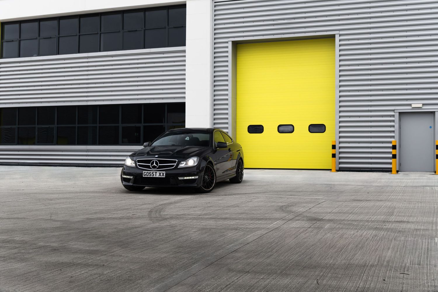 Black Mercedes-AMG C63 coupe parked in wide concrete yard in front of large yellow garage doors.