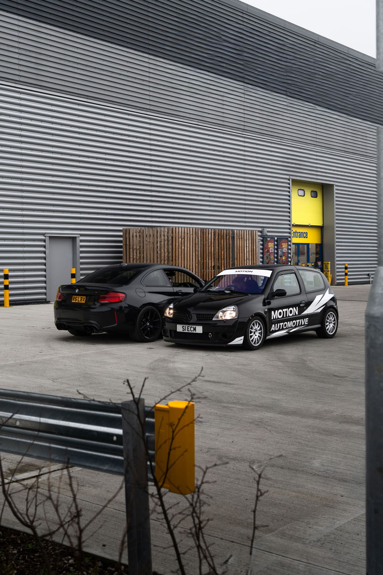 Two black modified cars parked outside Motion Automotive workshop with yellow roller doors.