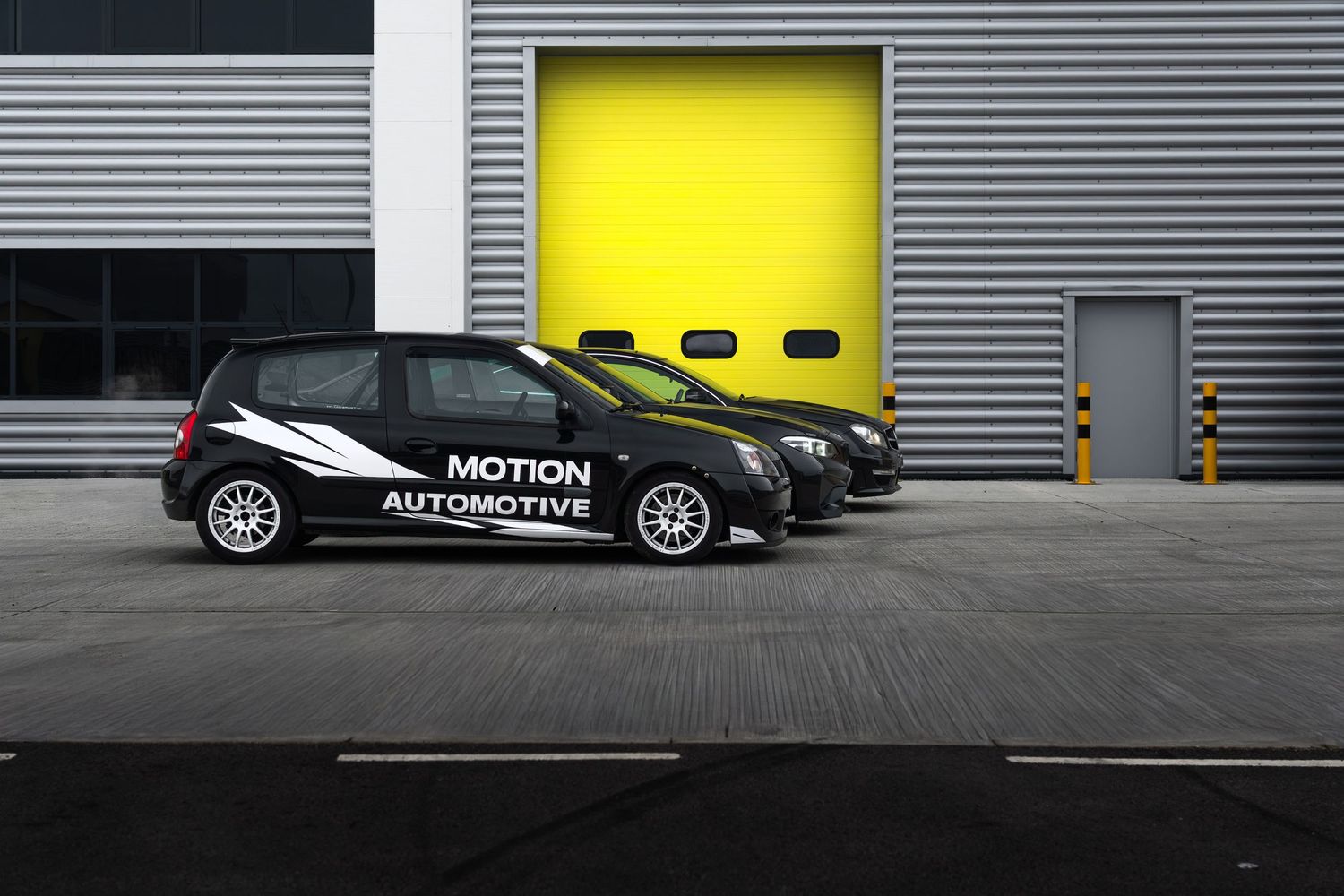 Three black Motion Automotive branded cars parked outside a industrial building with yellow roller doors.