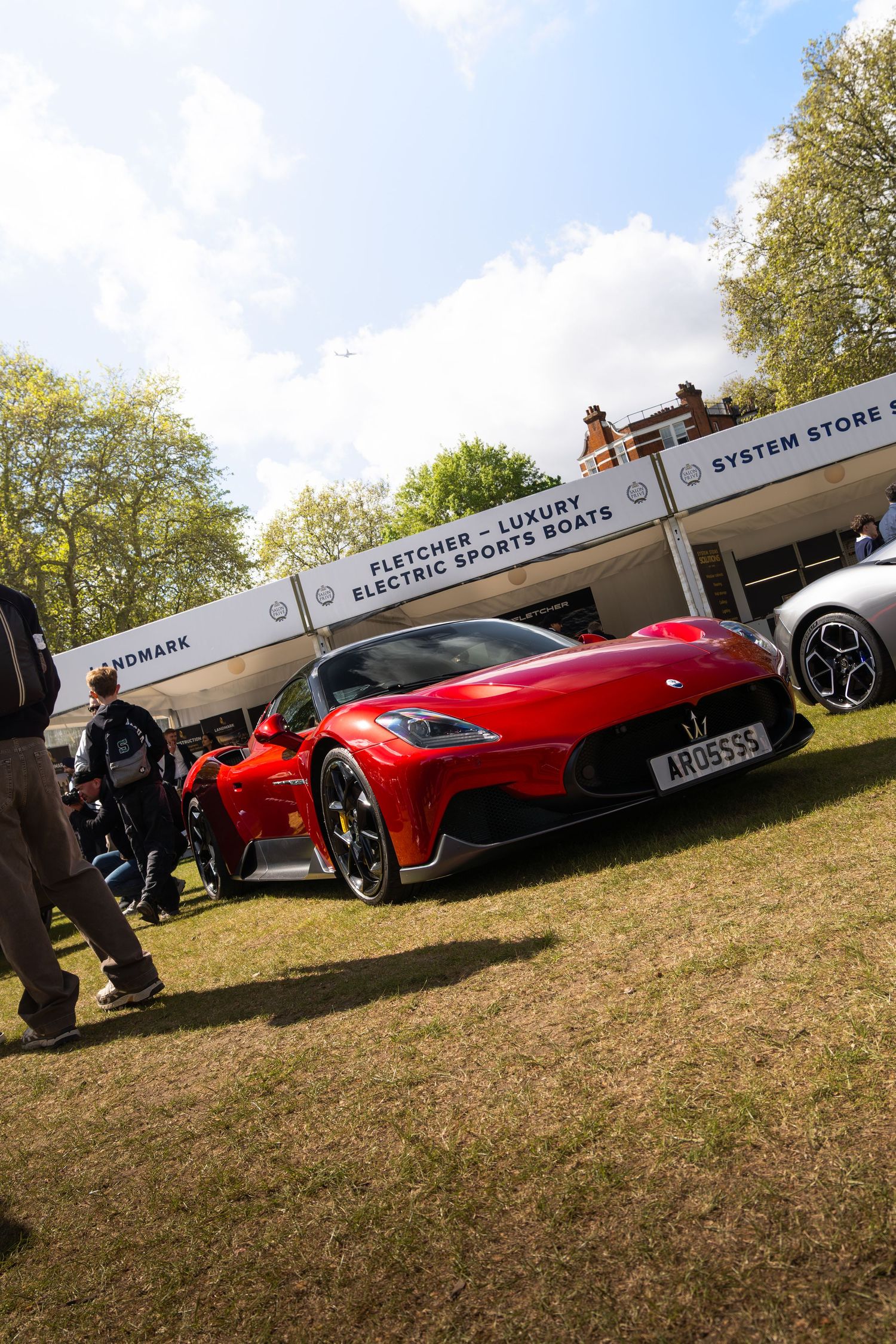 Red Maserati MC20 on display at an outdoor luxury car show under a partly cloudy sky.