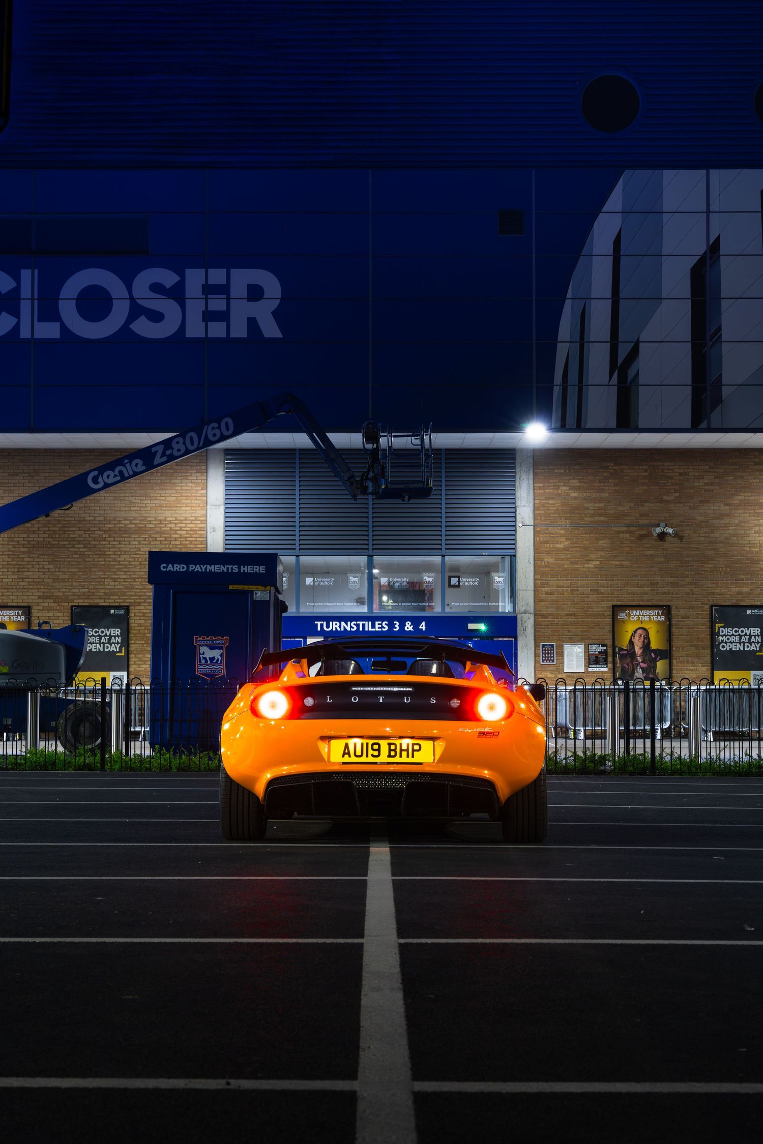 Orange Lotus Elise AU19BHP with brake lights glowing outside stadium Turnstiles 3 and 4 entrance at night.