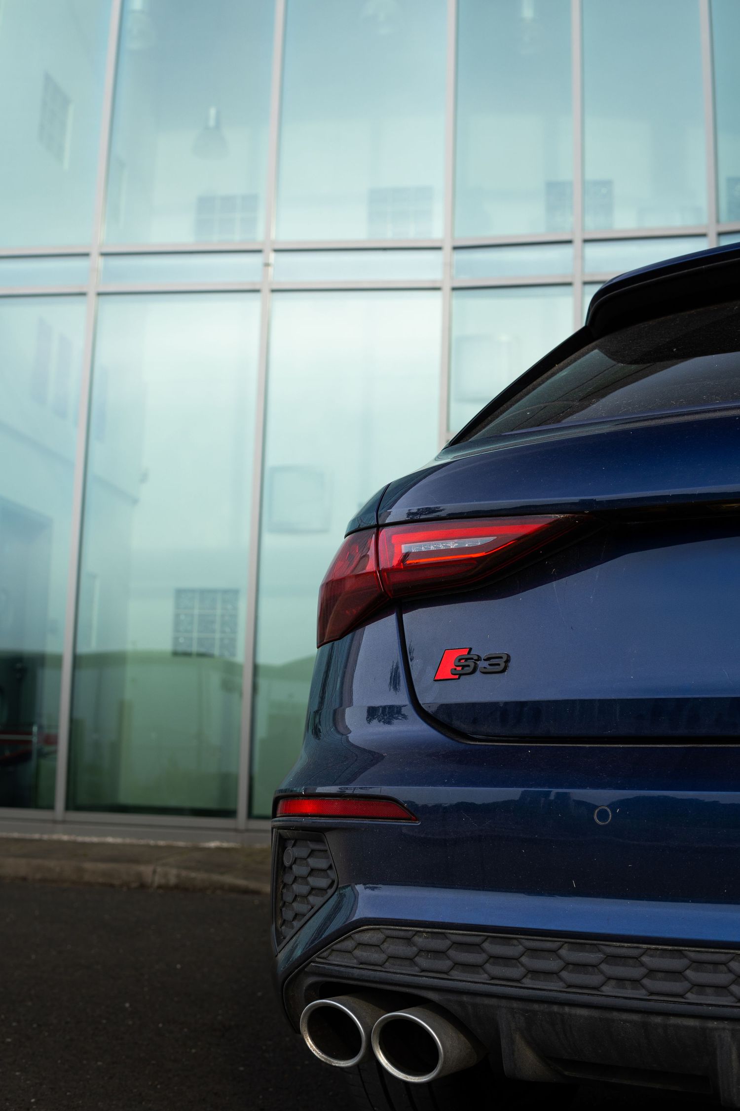 Close-up of blue Audi S3 rear badge and quad exhaust tips parked against a glass building.