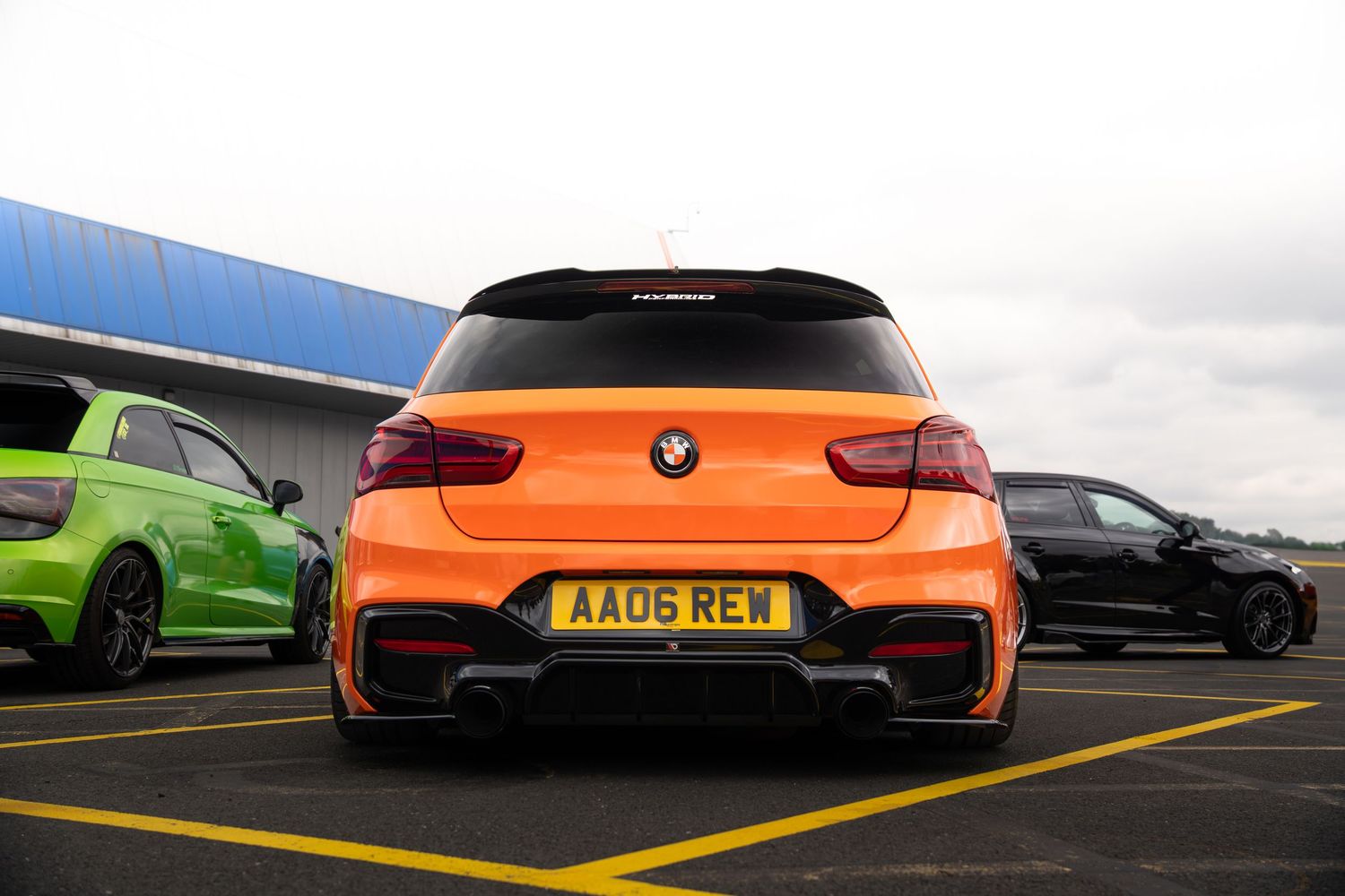 Rear view of a custom orange BMW with black spoiler and AA06 REW license plate in a parking lot.