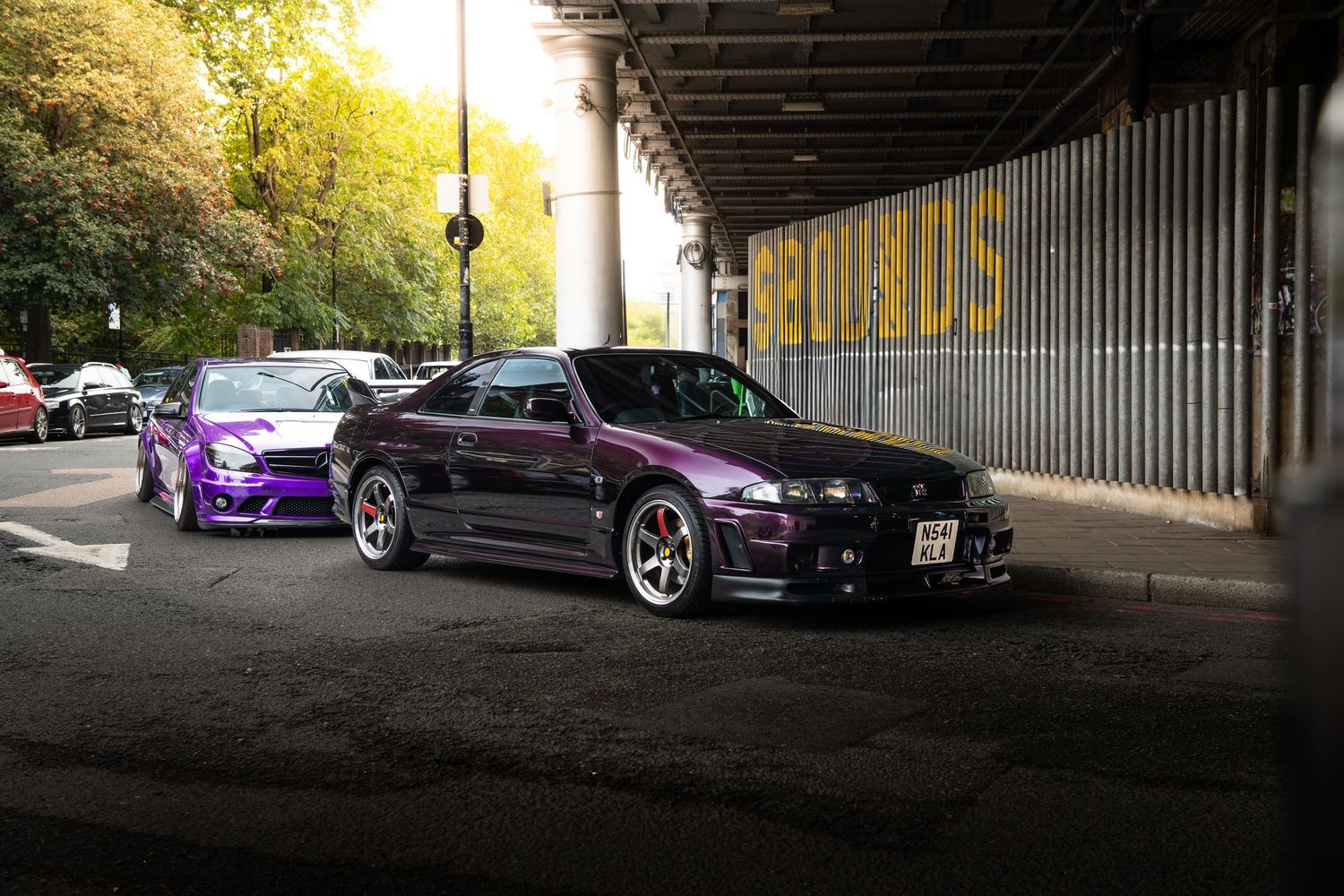 Two modified Nissan Skyline R34s in purple parked under a bridge with golden autumn trees.