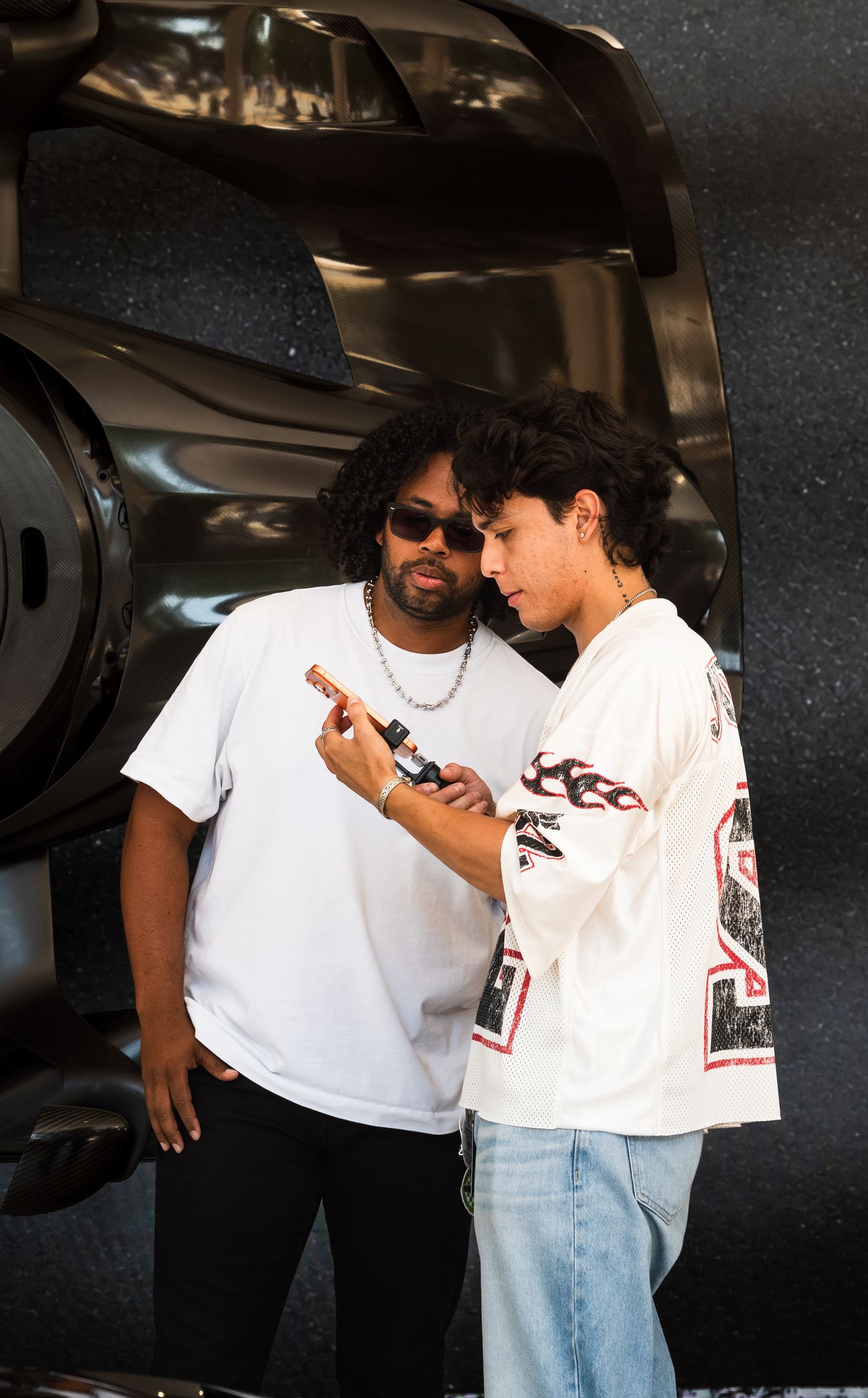 Two young men in white t-shirts examine a phone near a dark luxury car outdoors.