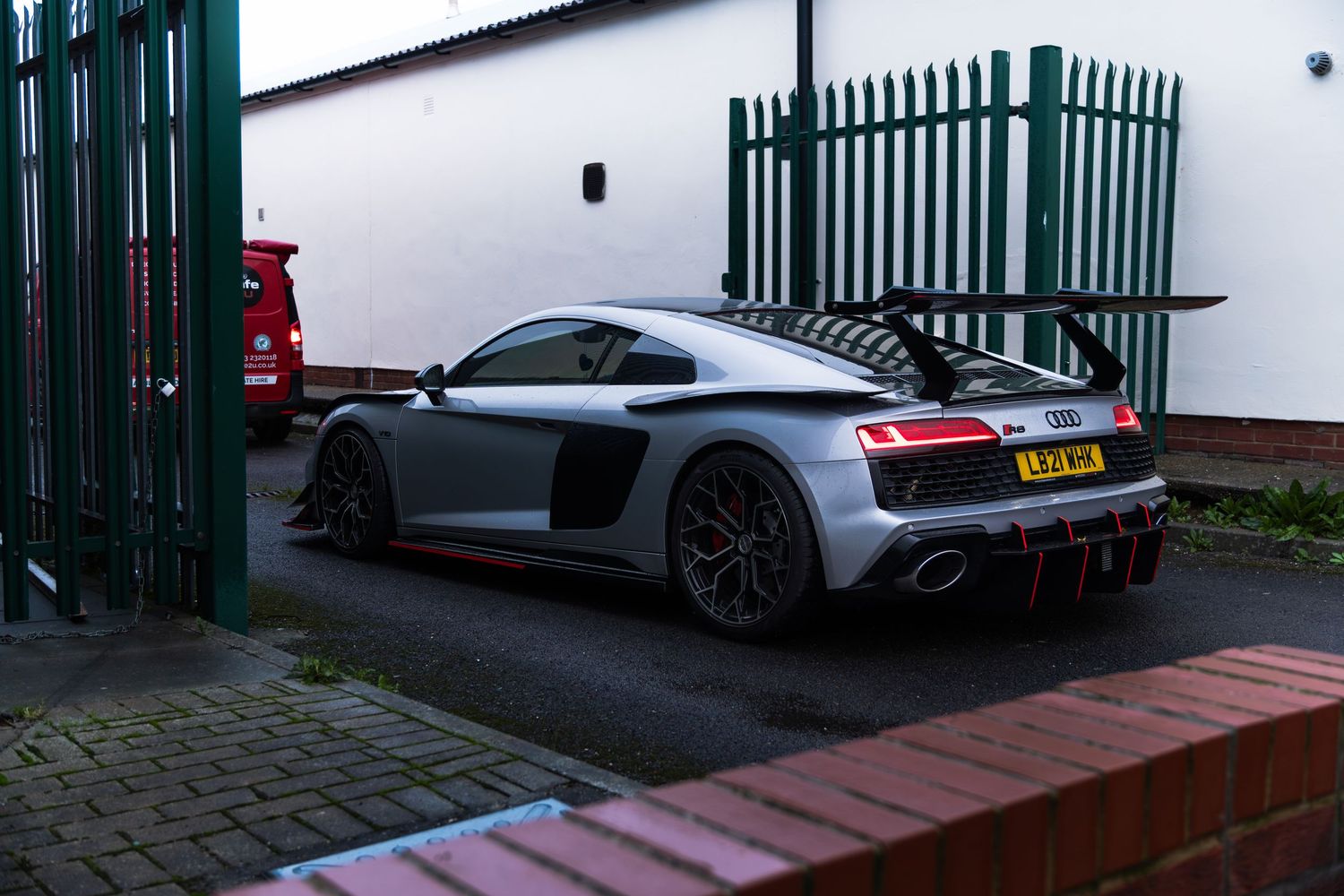 Modified silver Audi R8 GT3 with large rear wing in industrial yard.